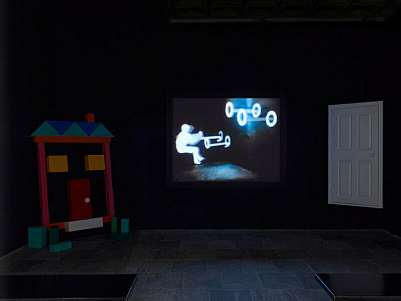 A darkened gallery with an art installation featuring a video projection, colorful house and white door.