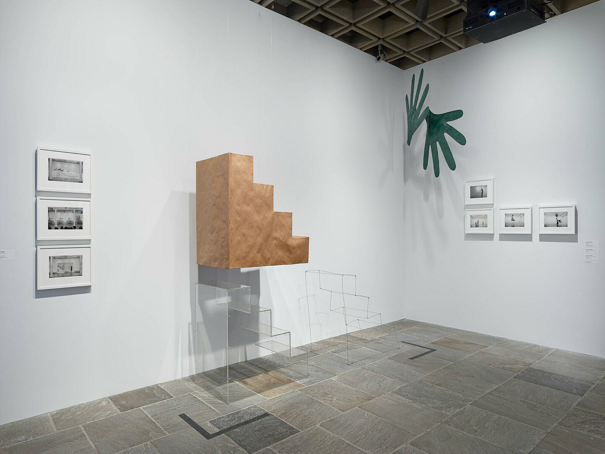 Art installation with photography on the walls, green hands in the corner and a wooden block of stairs.