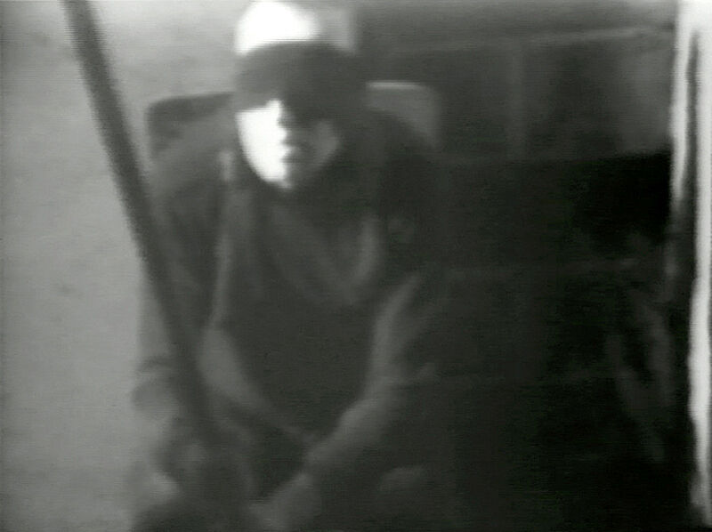 A black and white video still of a man with a blind fold on.
