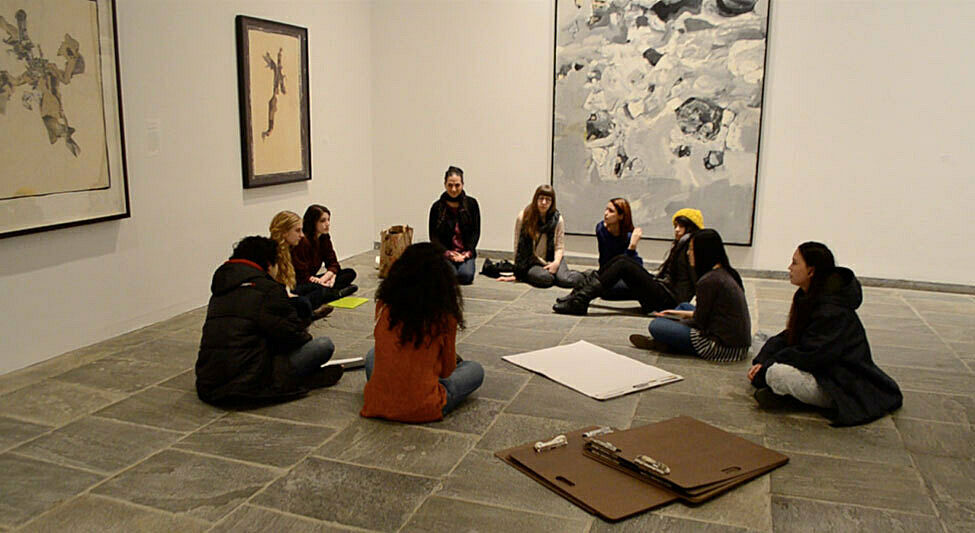 Students sit on a gallery floor and talk in a circle.