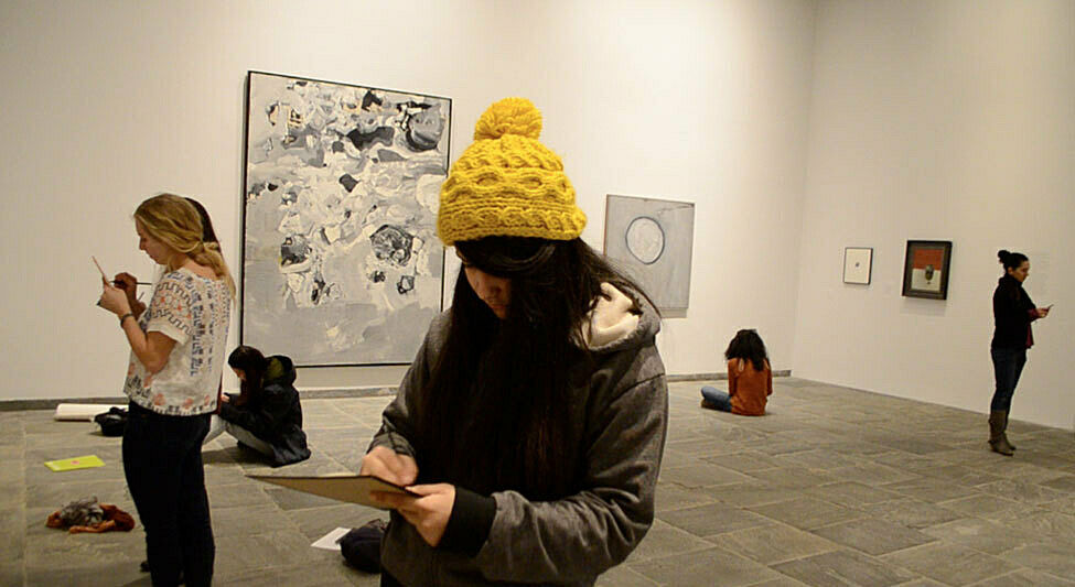 Students stand in front of artworks in the gallery as they sketch on paper. 