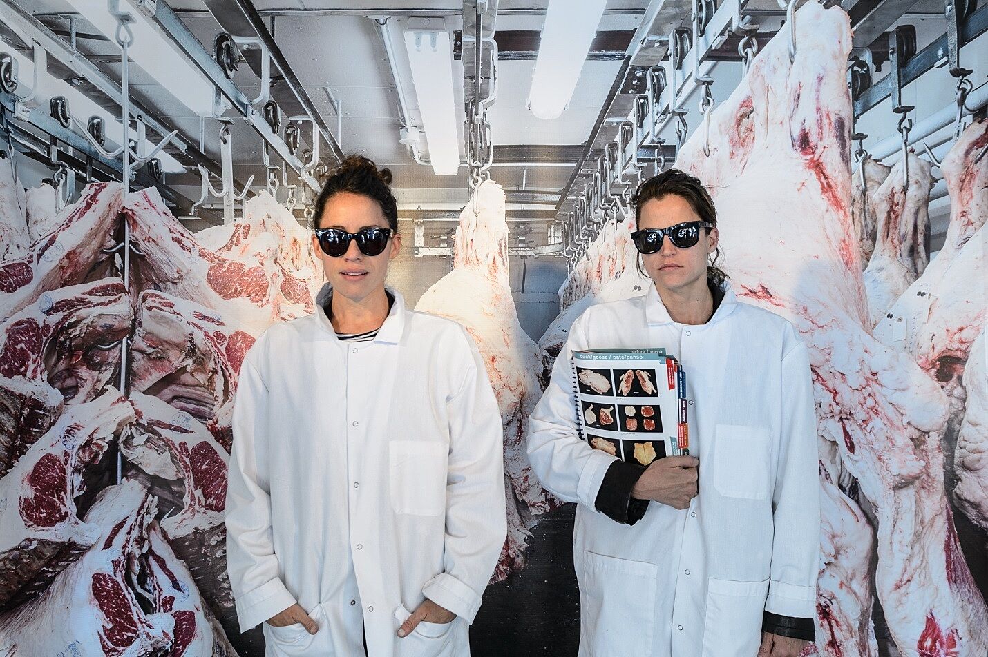 Two women wear white coats in front of a photo of a meatpacking plant.