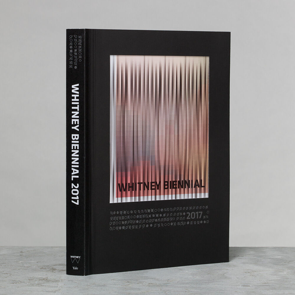 A black Whitney Biennial 2017 catalogue with a blurred vertical-striped cover image standing upright.