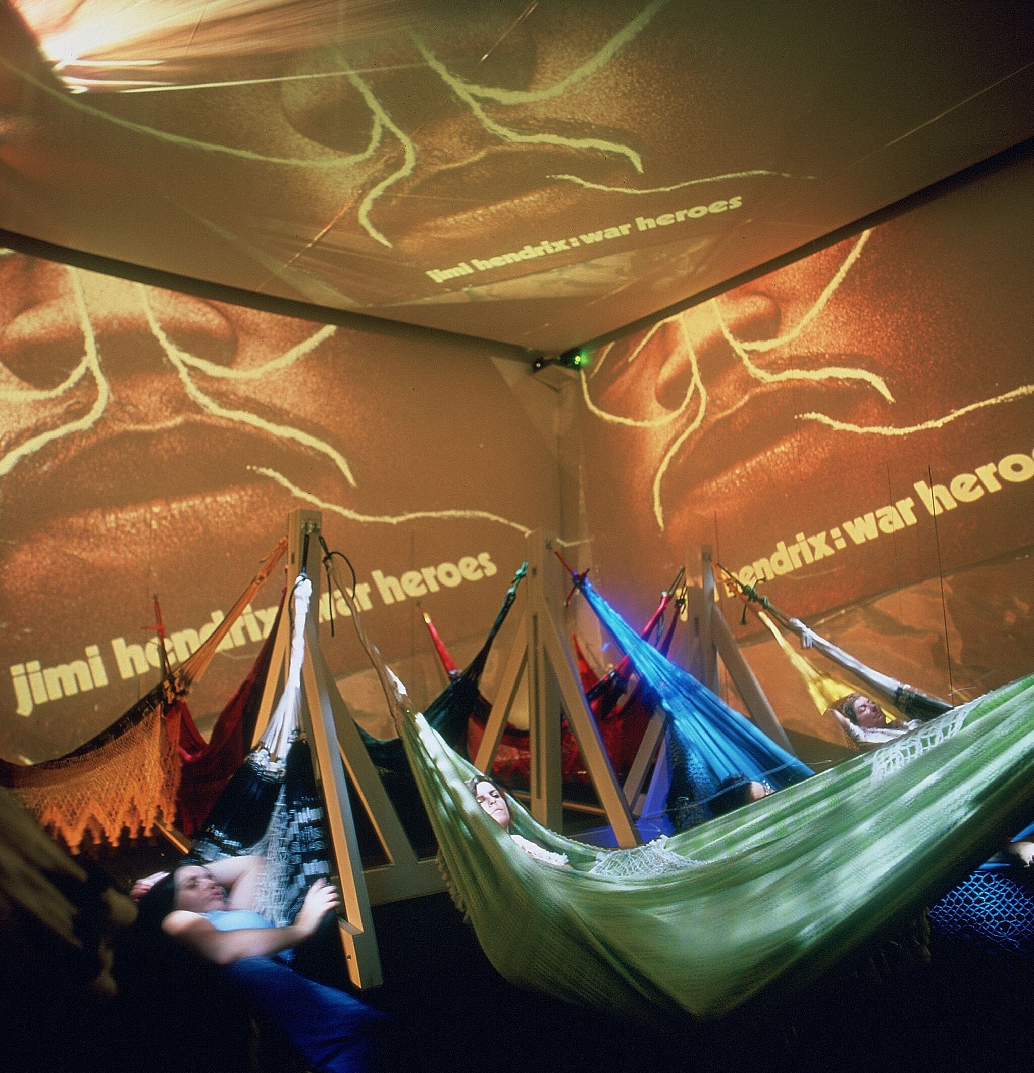 People lay in multicolored hammocks with projections on the walls behind them.
