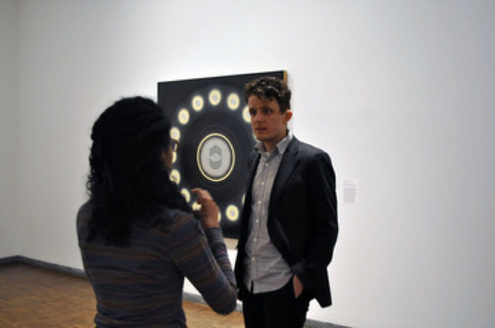 Two people in a gallery speaking to each other in front of an artwork on the wall.