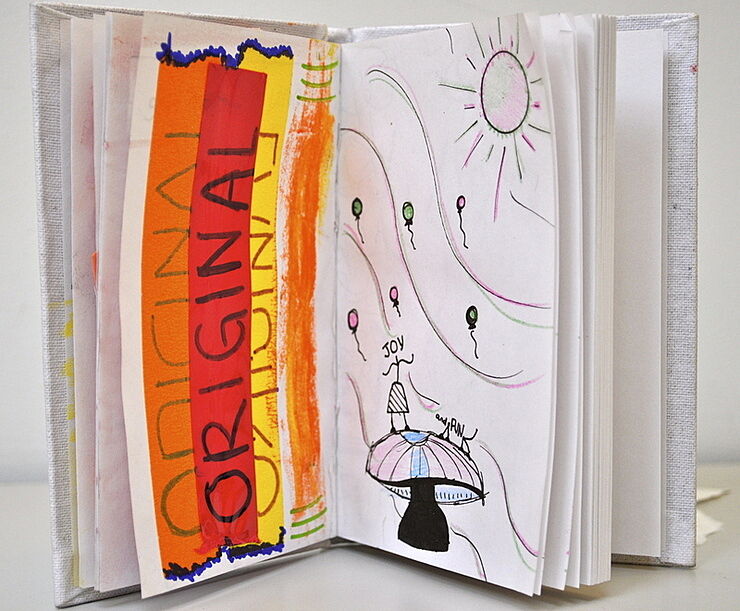 Open sketchbook with colorful drawings and text. Left page has "ORIGINAL" in bold letters, right page shows a sun, balloons, and a figure with "JOY" written.