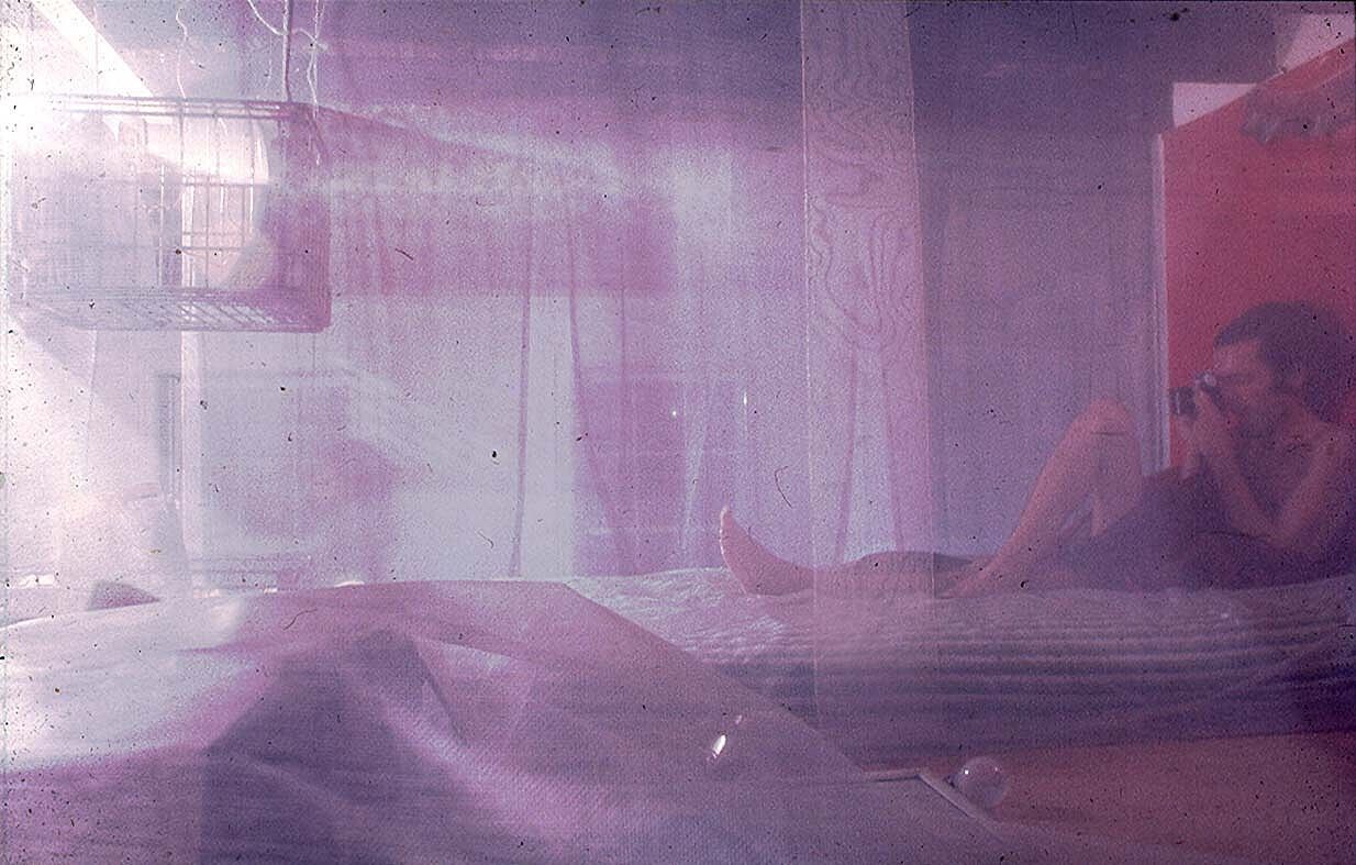 A man laying down and taking a photo is seen behind a purple tinted translucent fabric. 