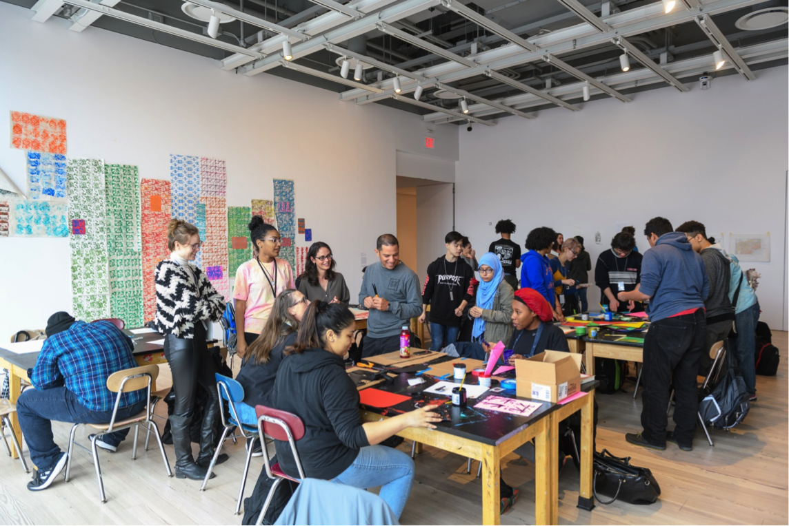 A group of people engaged in an art workshop, surrounded by colorful prints on the wall and working at tables with art supplies.