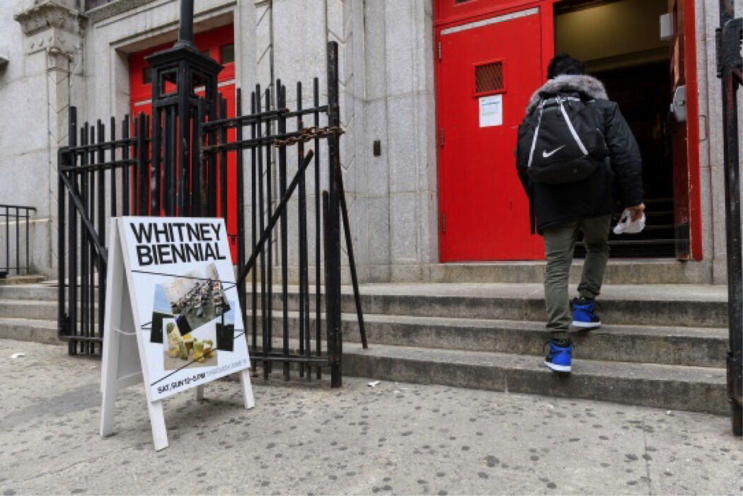 A person wearing a backpack walks up steps into a building beside a Whitney Biennial sign.