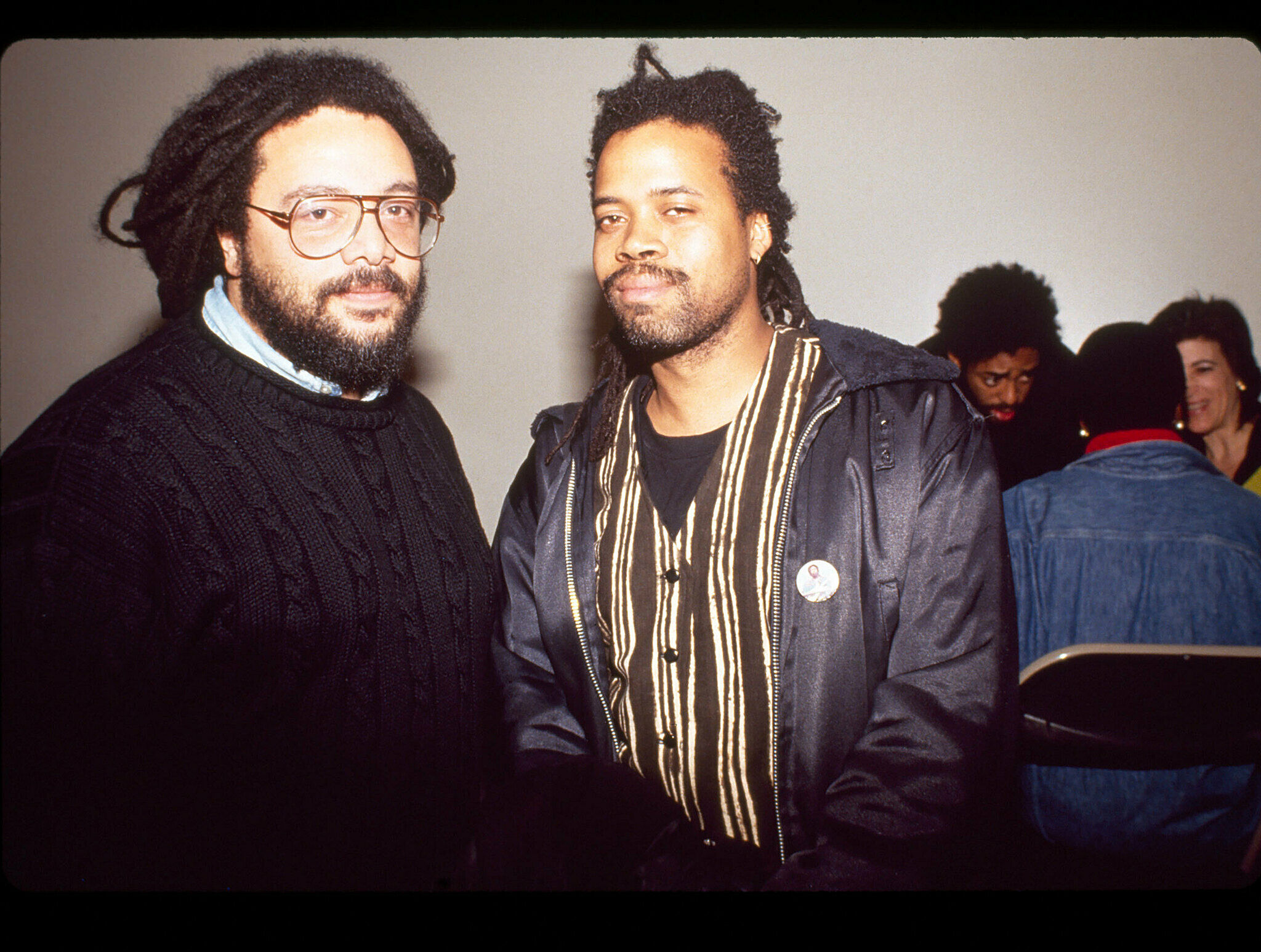 Two men with dreadlocks smiling at the camera, one in a black sweater, the other in a leather jacket, with people in the background.