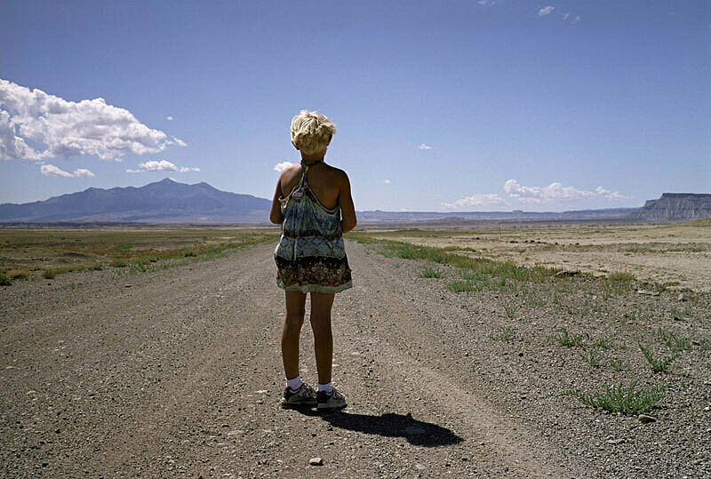 A small person stands on a dirt road in the desert, facing away from the camera.