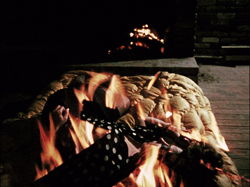 Still from film HOLD ME WHILE I’M NAKED. Image depicts a bed with blankets in the foreground and a fireplace in the background.
