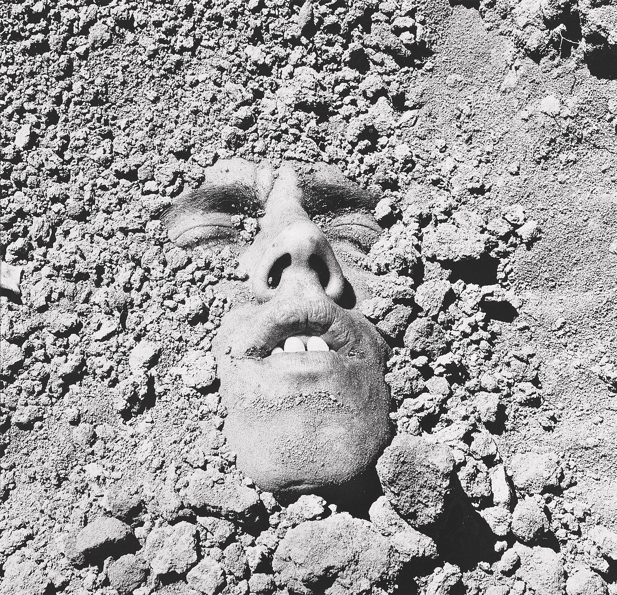 A photograph of a man's face emerging from gravel. 