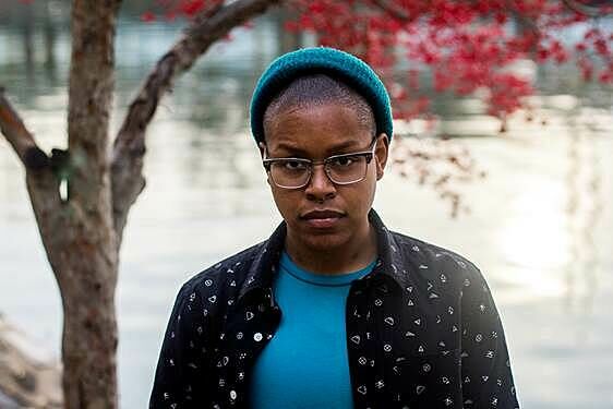 Cyree, a queer black male, is photographed wearing a bright blue shirt and beanie hat, glasses, and a black and white patterned sleeved shirt overtop. He's standing in front of a lake with a large red tree