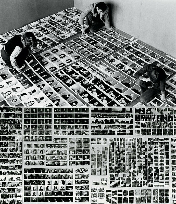 People looking at many black and white photographs on the floor. 
