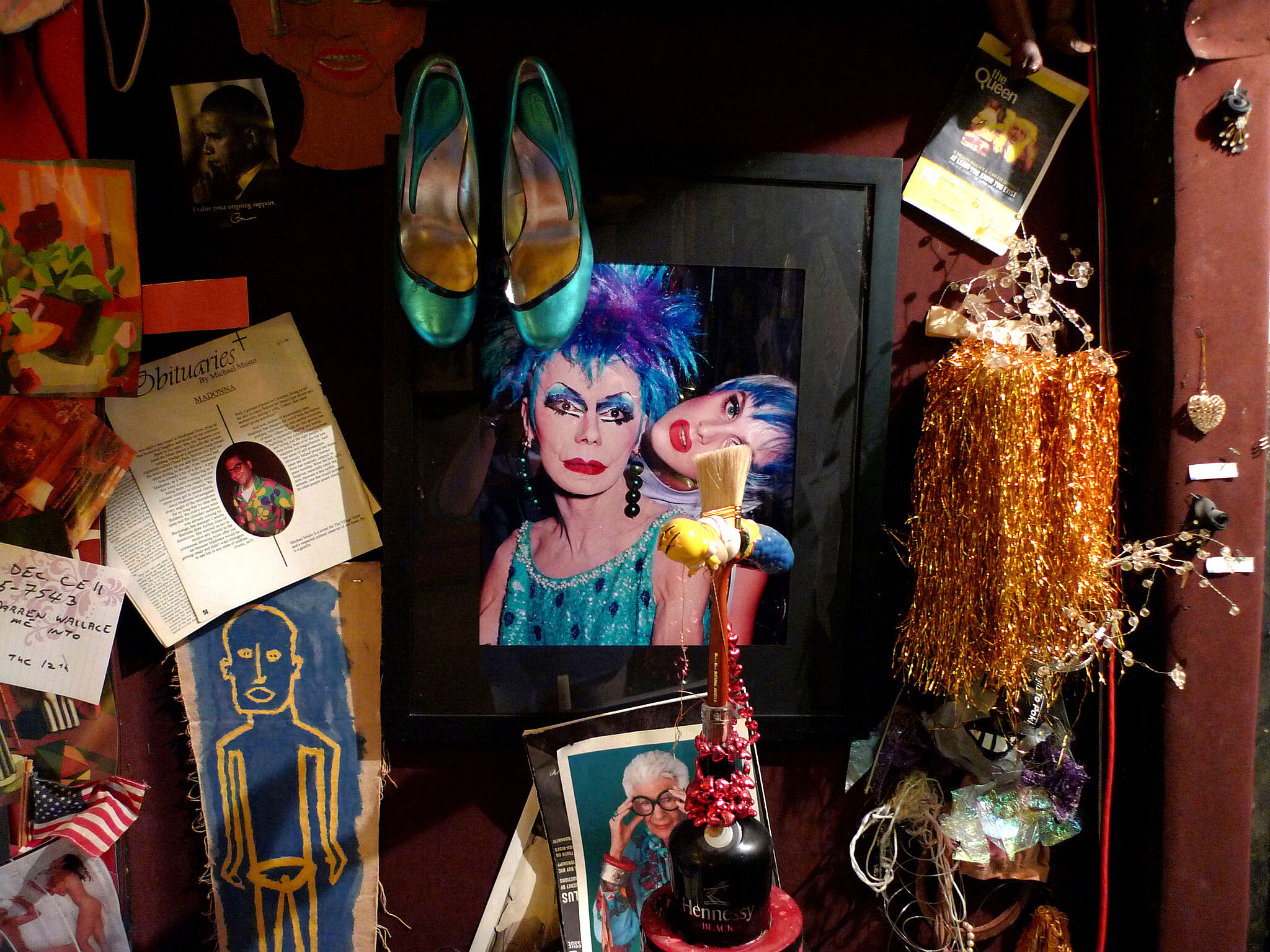 Eclectic memorabilia wall with a framed photo of a woman in drag, hanging teal shoes, and various trinkets.