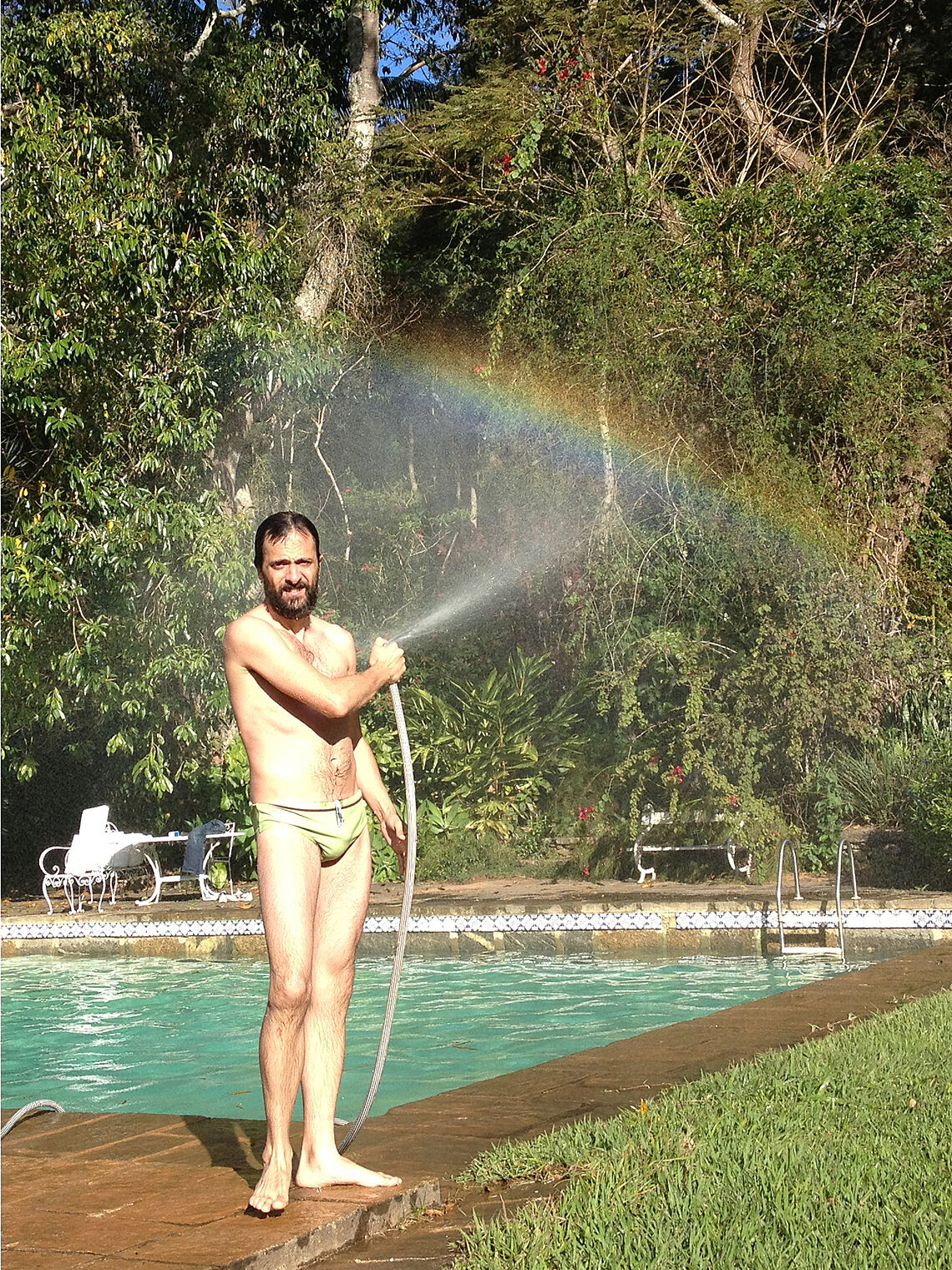 A man standing by a swimming pool playing with a sprinkler pipe. 
