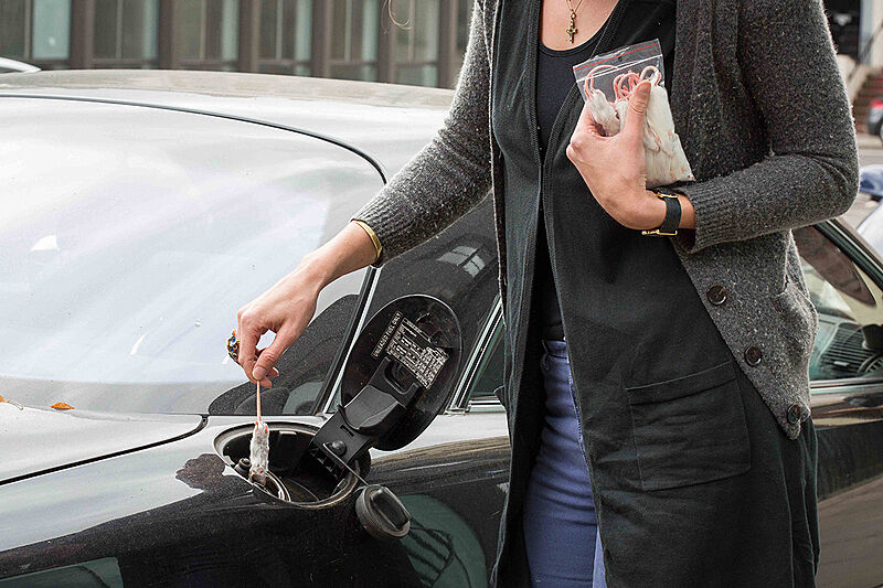 A woman holding a bag of lab rats and about to put one into the fuel hole of a car. 