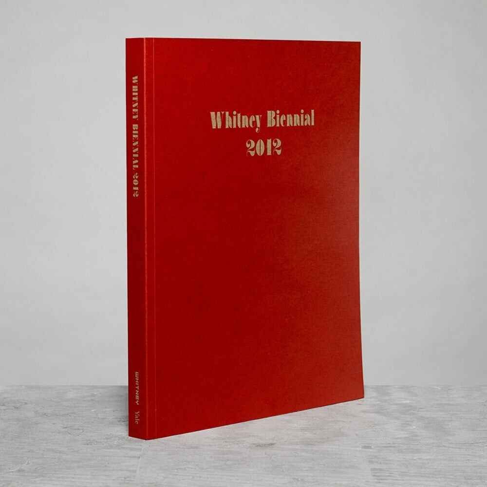 Red book standing upright titled "Whitney Biennial 2012" on a plain surface.
