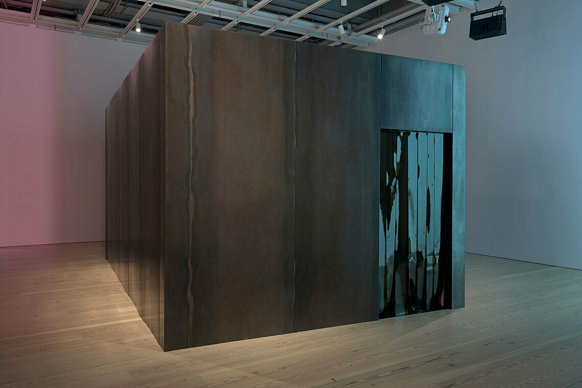 Large metallic cube with a reflective slit in a gallery space, under soft lighting.