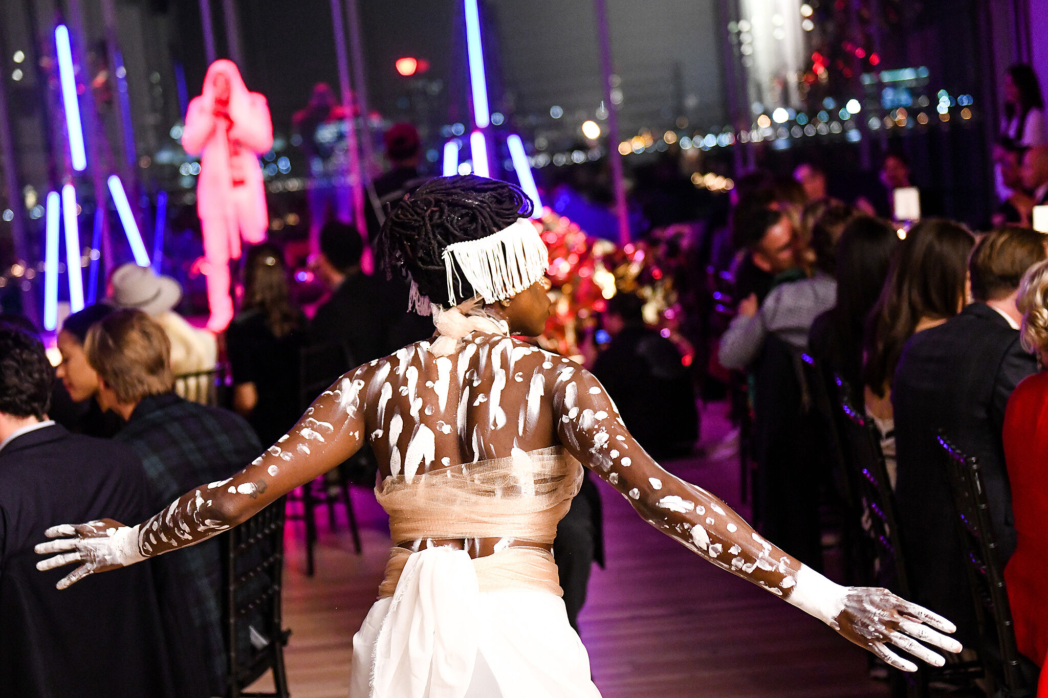 Person with painted skin and headpiece at a party with neon lights and cityscape in the background.