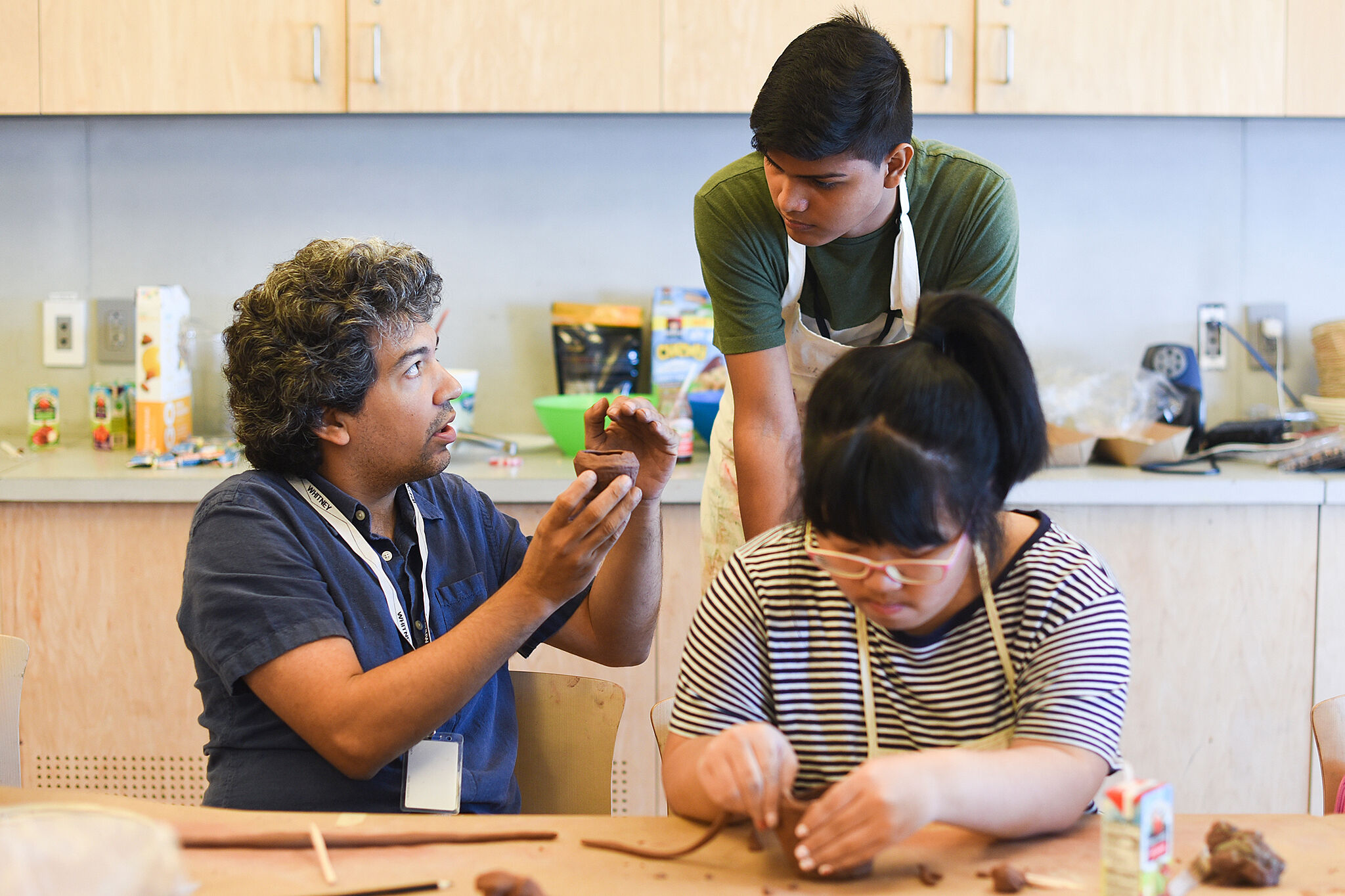 Instructor, Jorge González, speaks to two students. One student works with clay at a table, and the other stands by and listens.