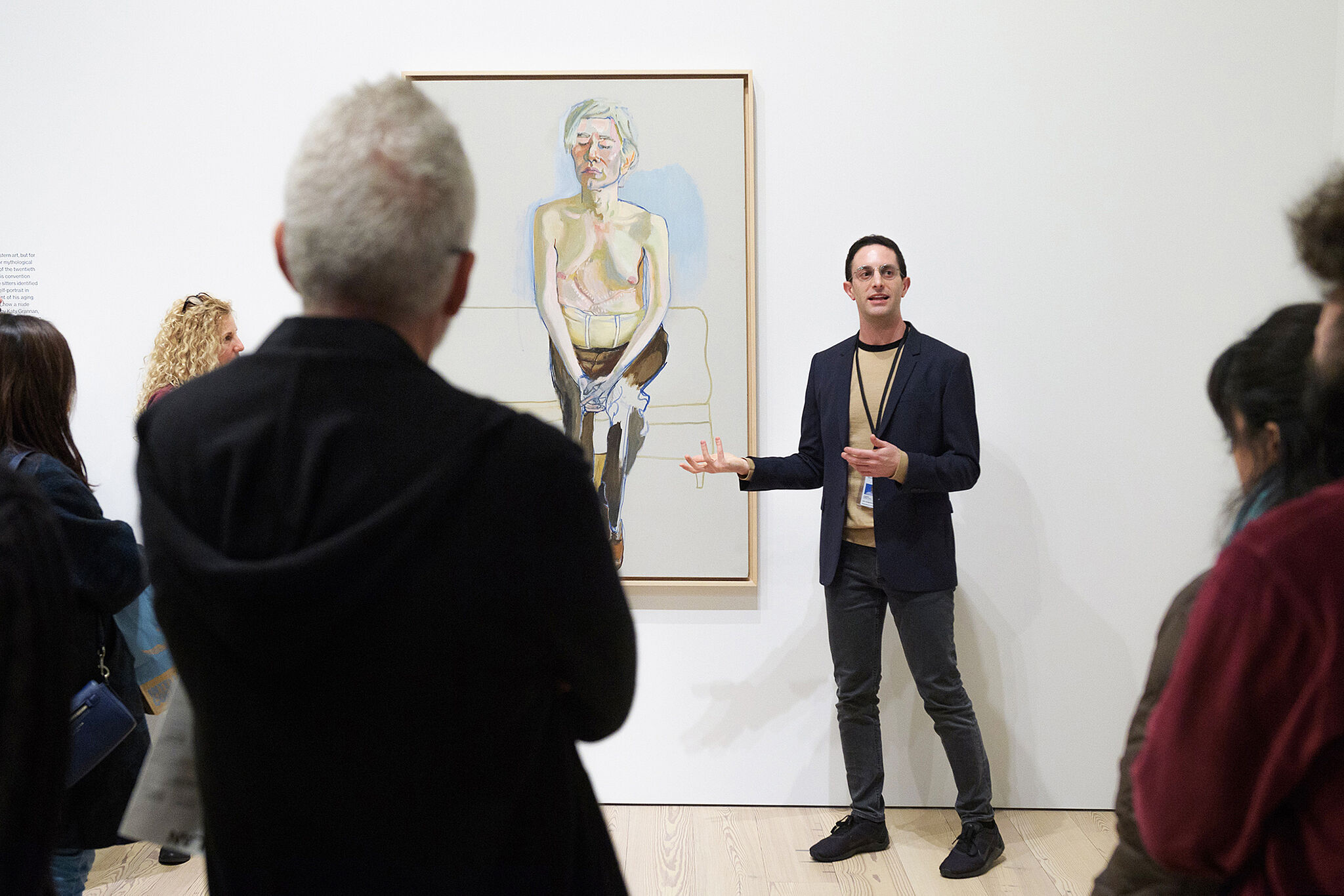 An educator stands in front of a work by Alice Neel, while students stand in the foreground.