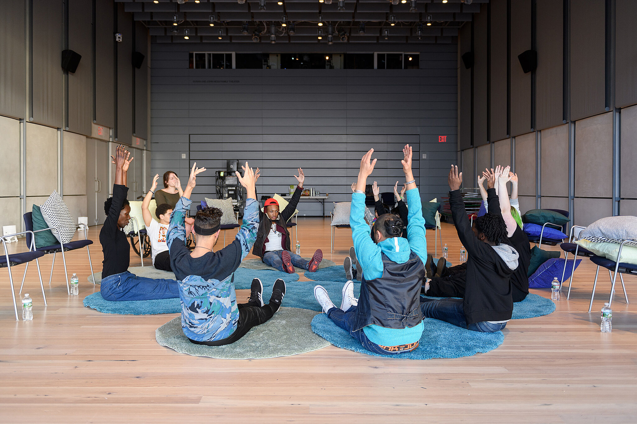 A photo of a group of people sitting on the floor in a circle, with hands raised above their heads.