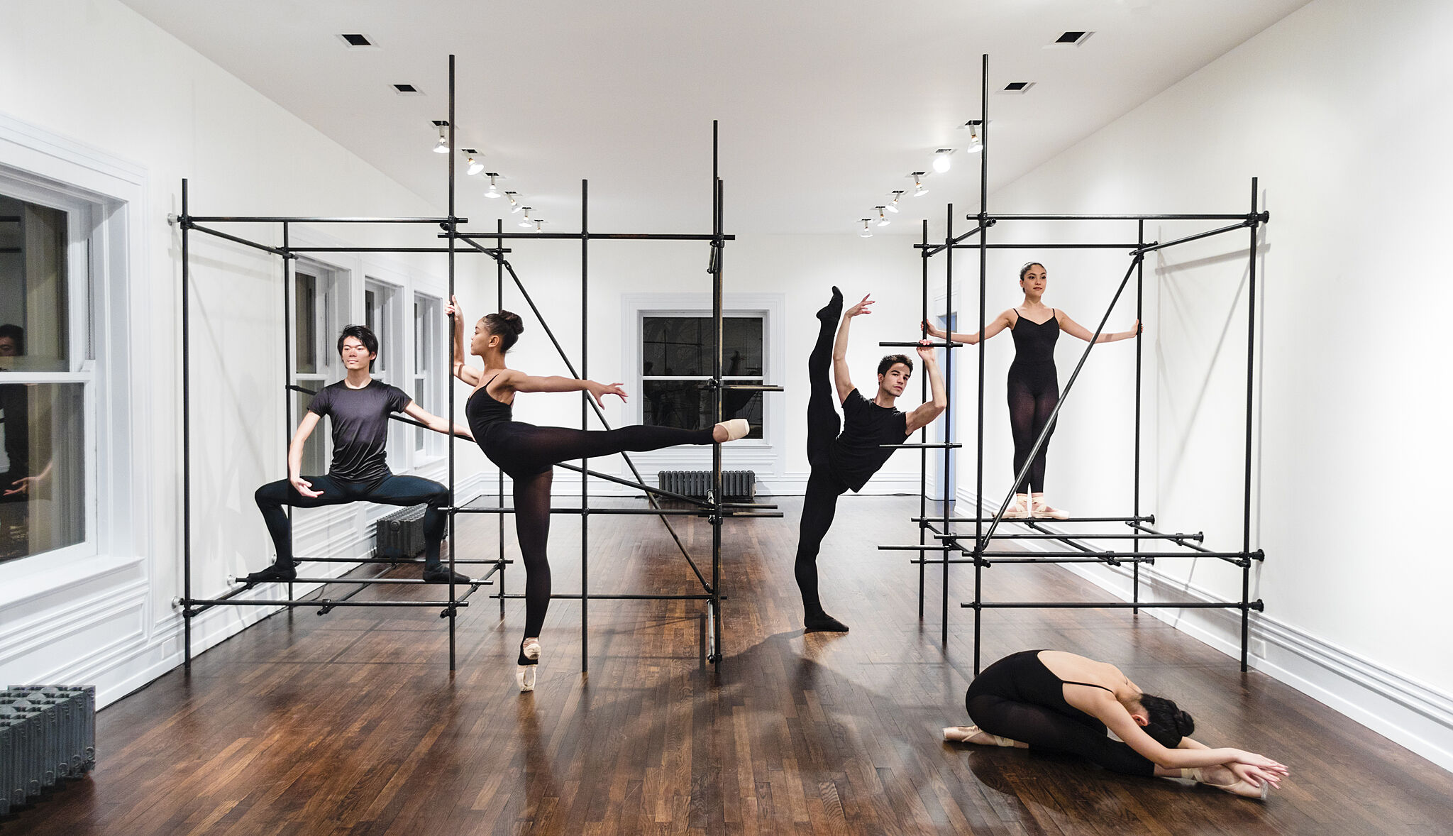 Four dancers pose in a rehearsal space with metal bar structures.