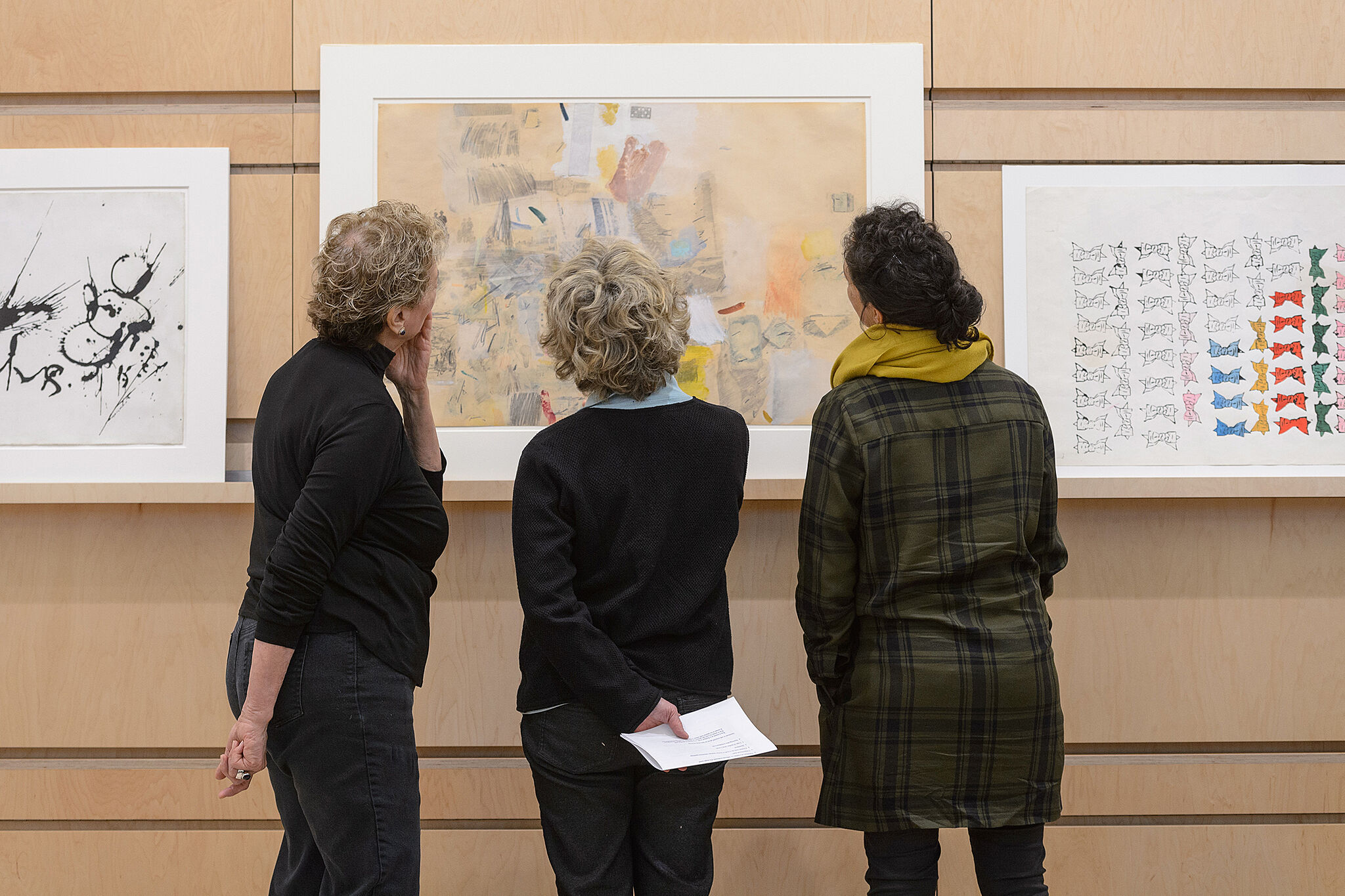 Three people examine an artwork, which hangs on a wall.