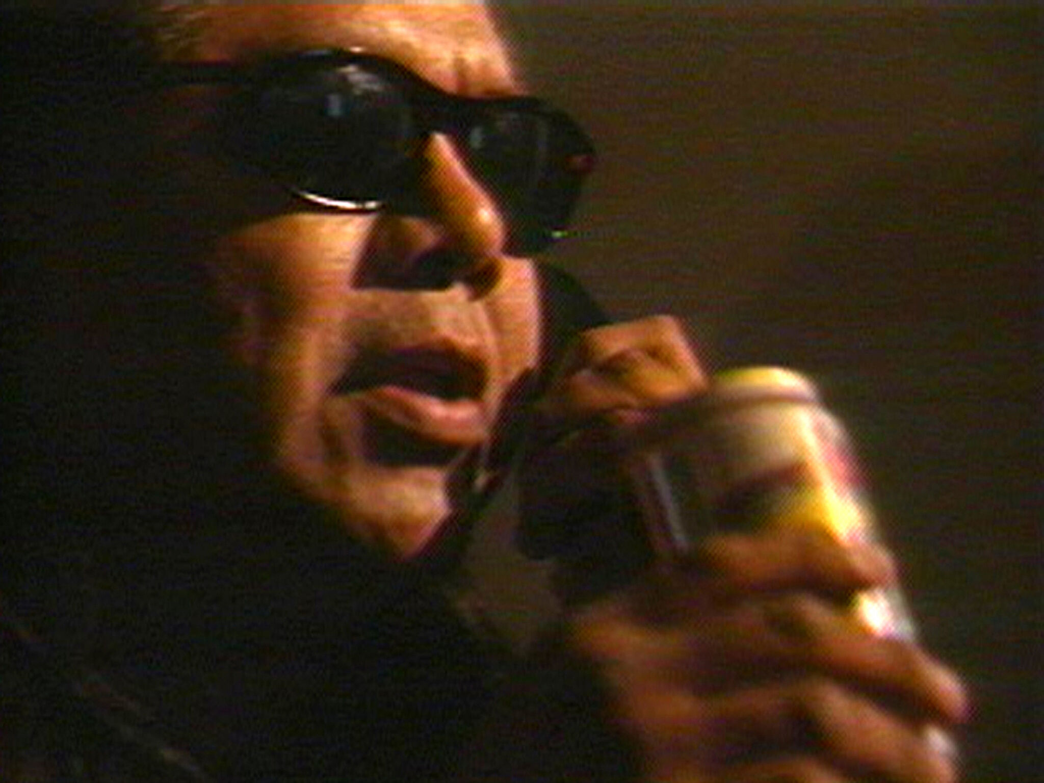 A close-up video still of a man wearing sunglasses, holding a can and a phone.