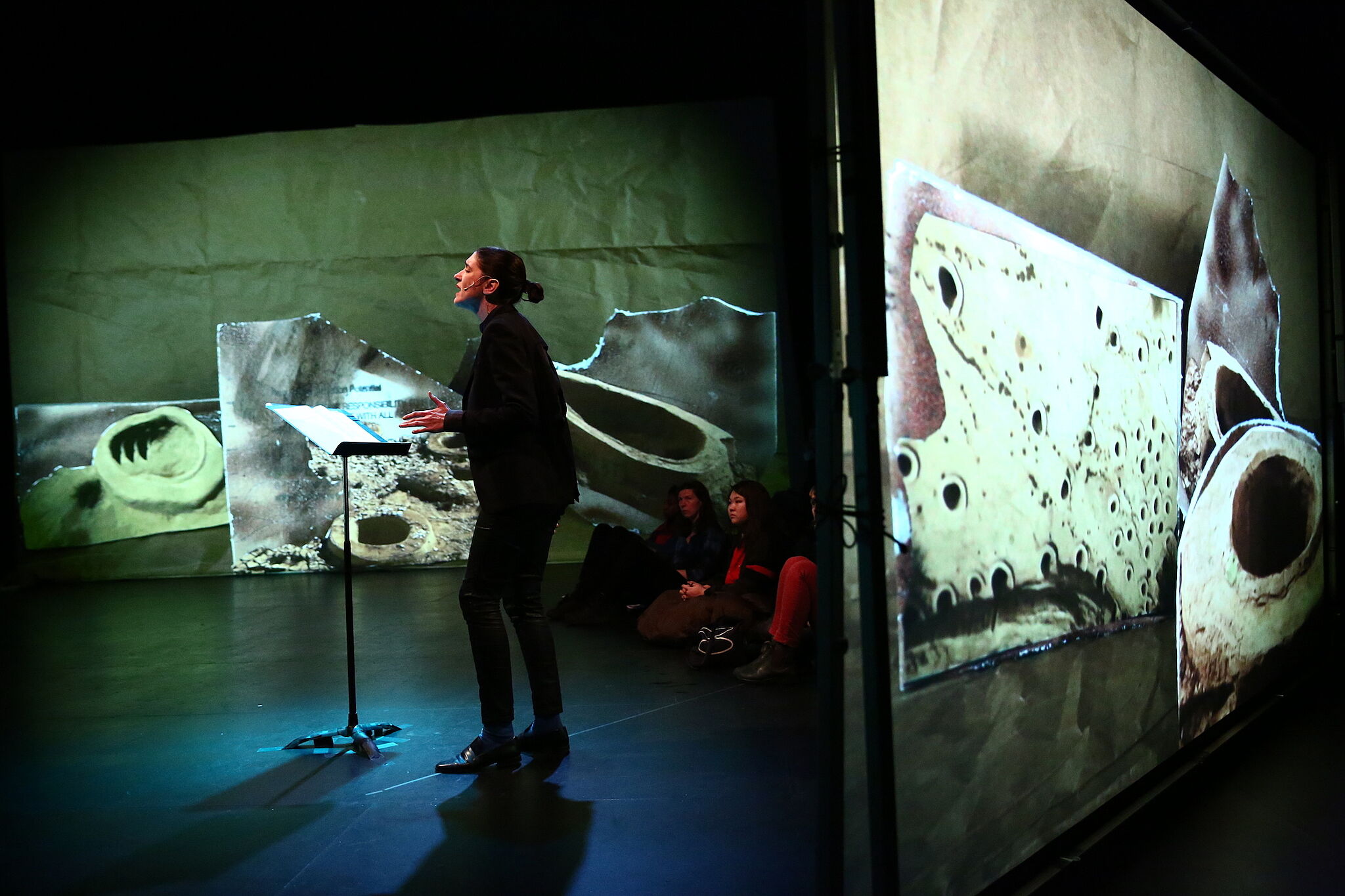 A photo of a person performing with a music stand surrounded by a multimedia performance. 