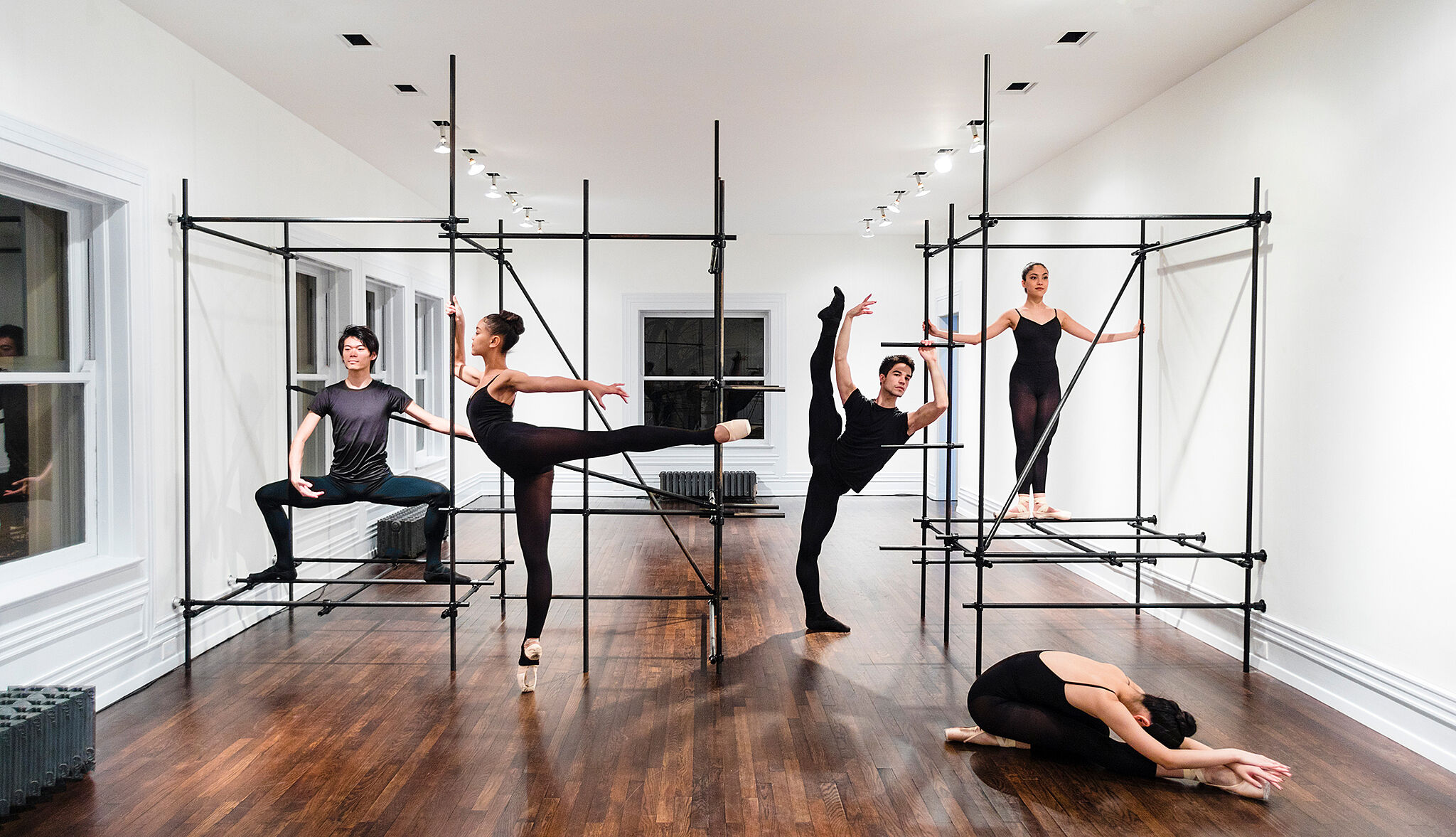 Four dancers pose in a rehearsal space with metal bar structures.