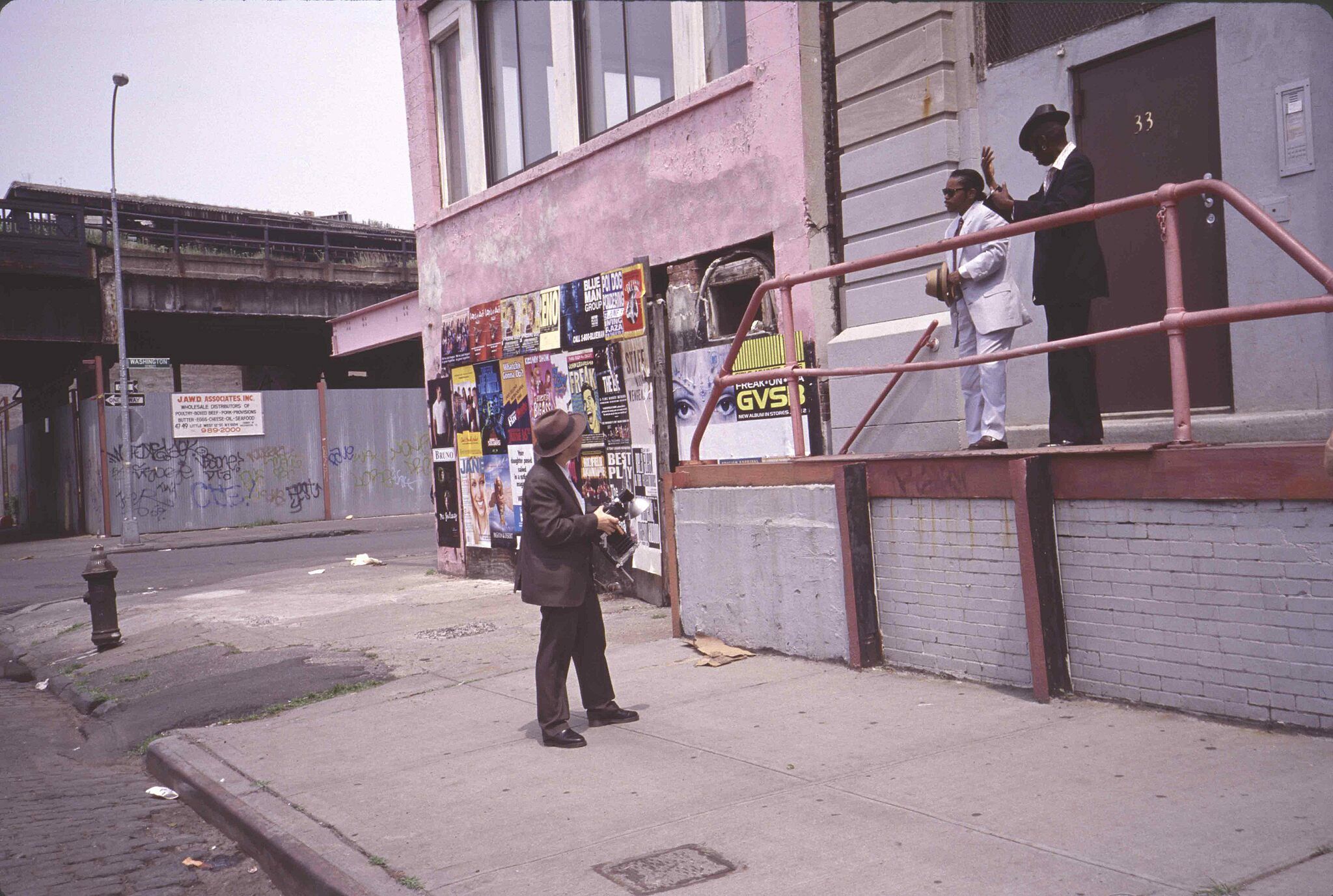 A photograph of two men on a platform looking at a man below them on the street holding a camera.