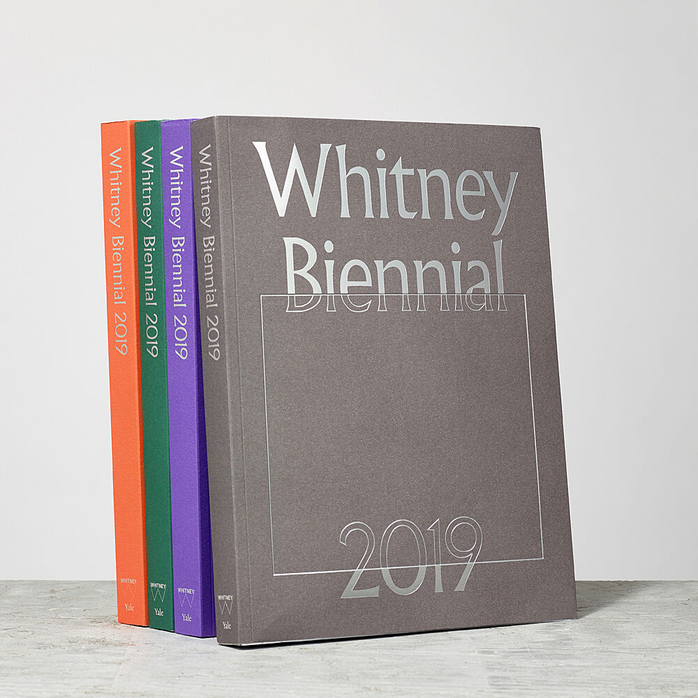 Whitney Biennial 2019 catalogues in four colors standing up in a row.