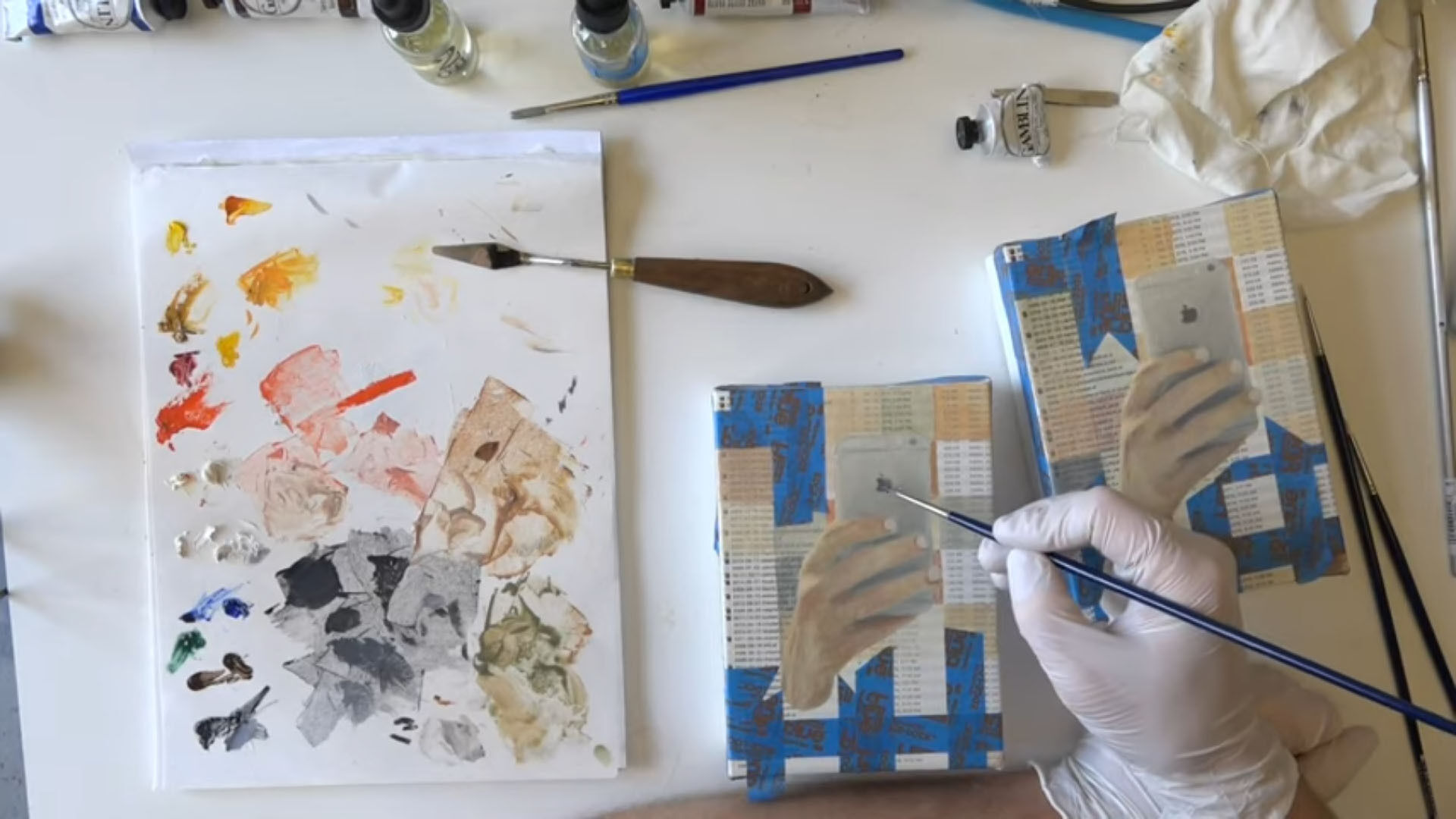 A hand painting a hand holding an iPhone, with a paper paint palette to the left side, viewed from above.