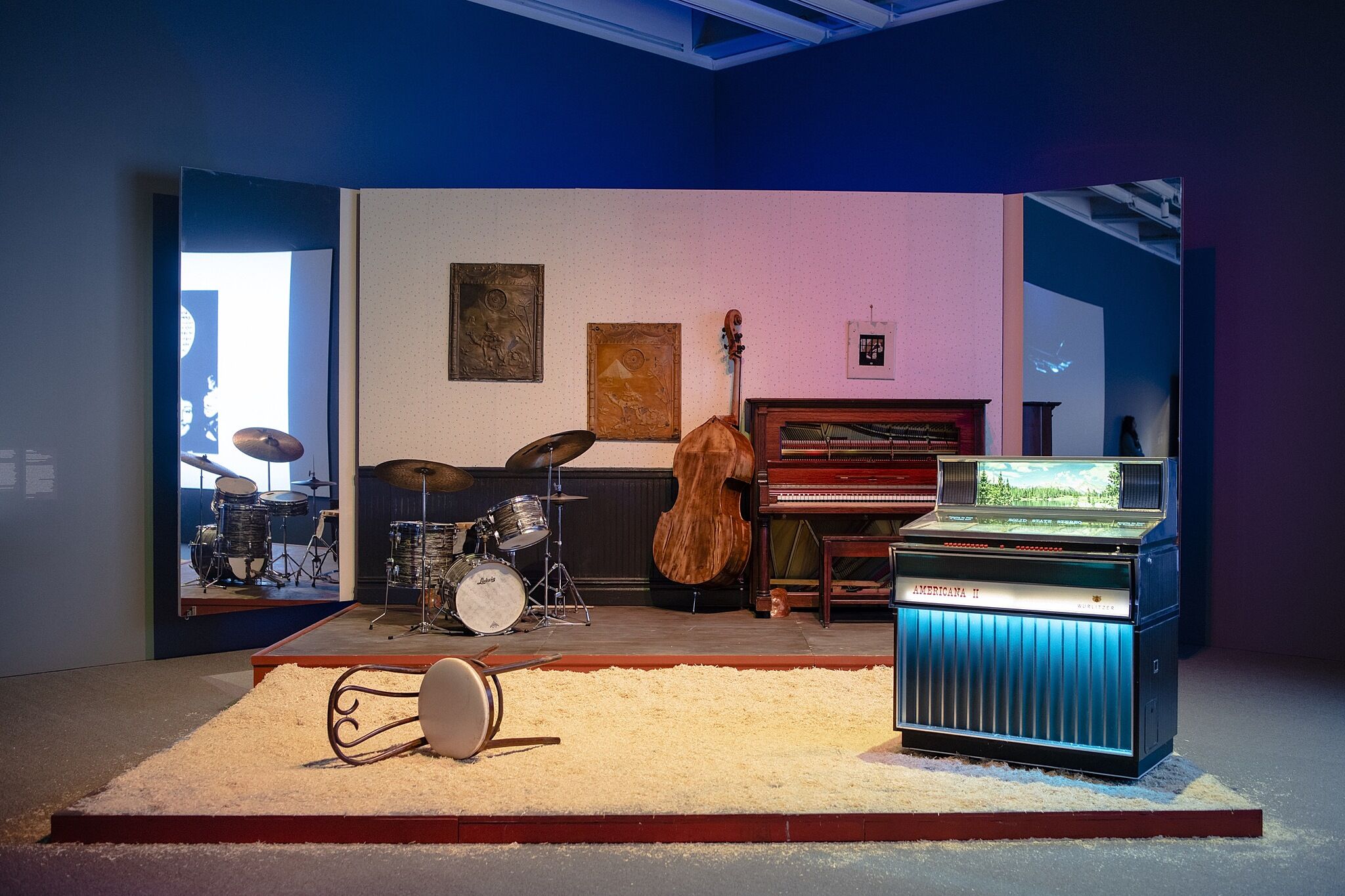 An image of a stage with drums, a bass, a piano, a jukebox, and an overturned chair