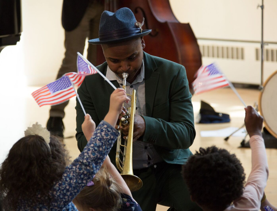 A photo of a person playing the trumpet surrounded by children waving American flags.