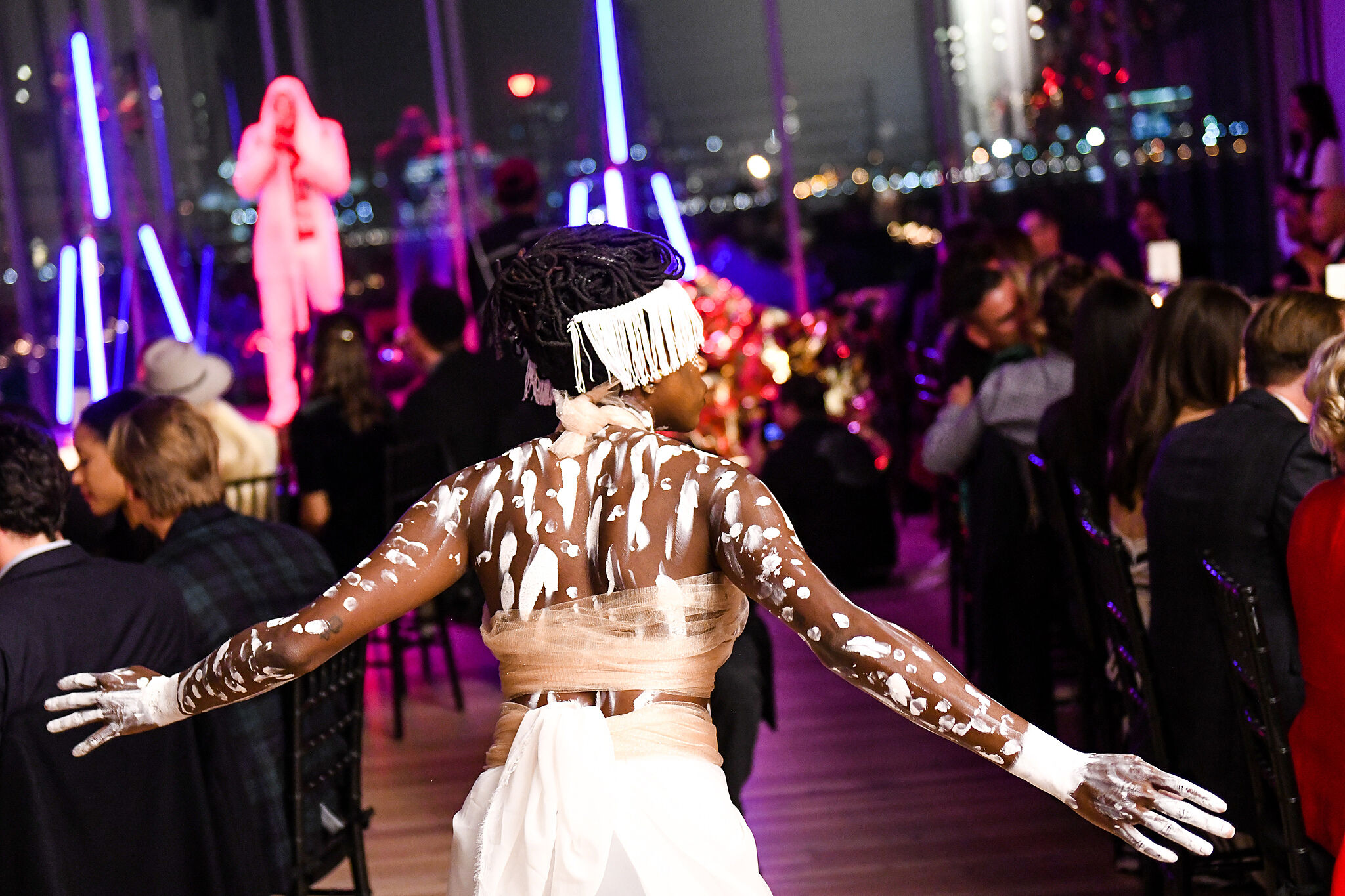An artist with white paint on their body walks through a crowded dining room.