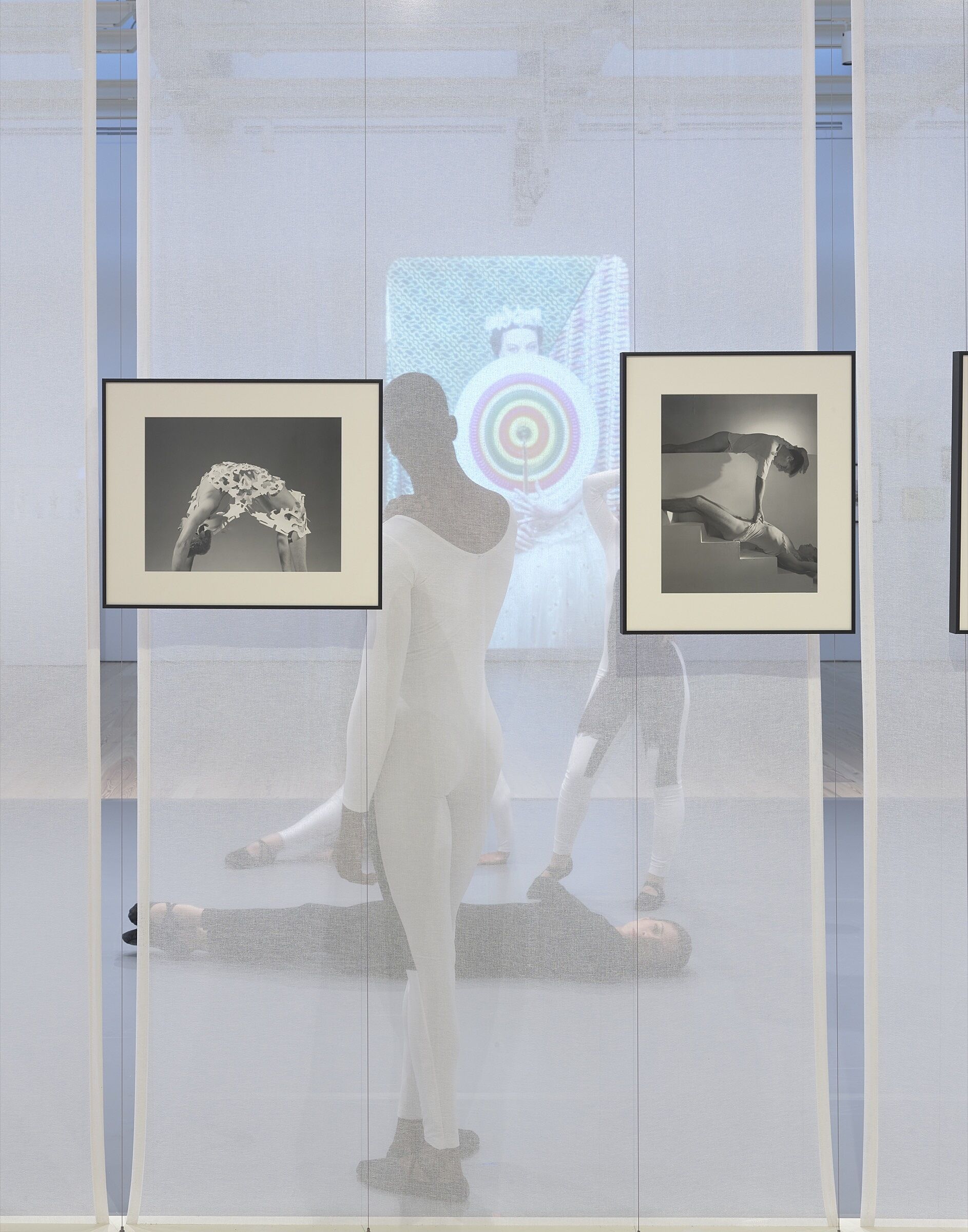 Four dancers perform behind a screen in a gallery space full of photos and video.