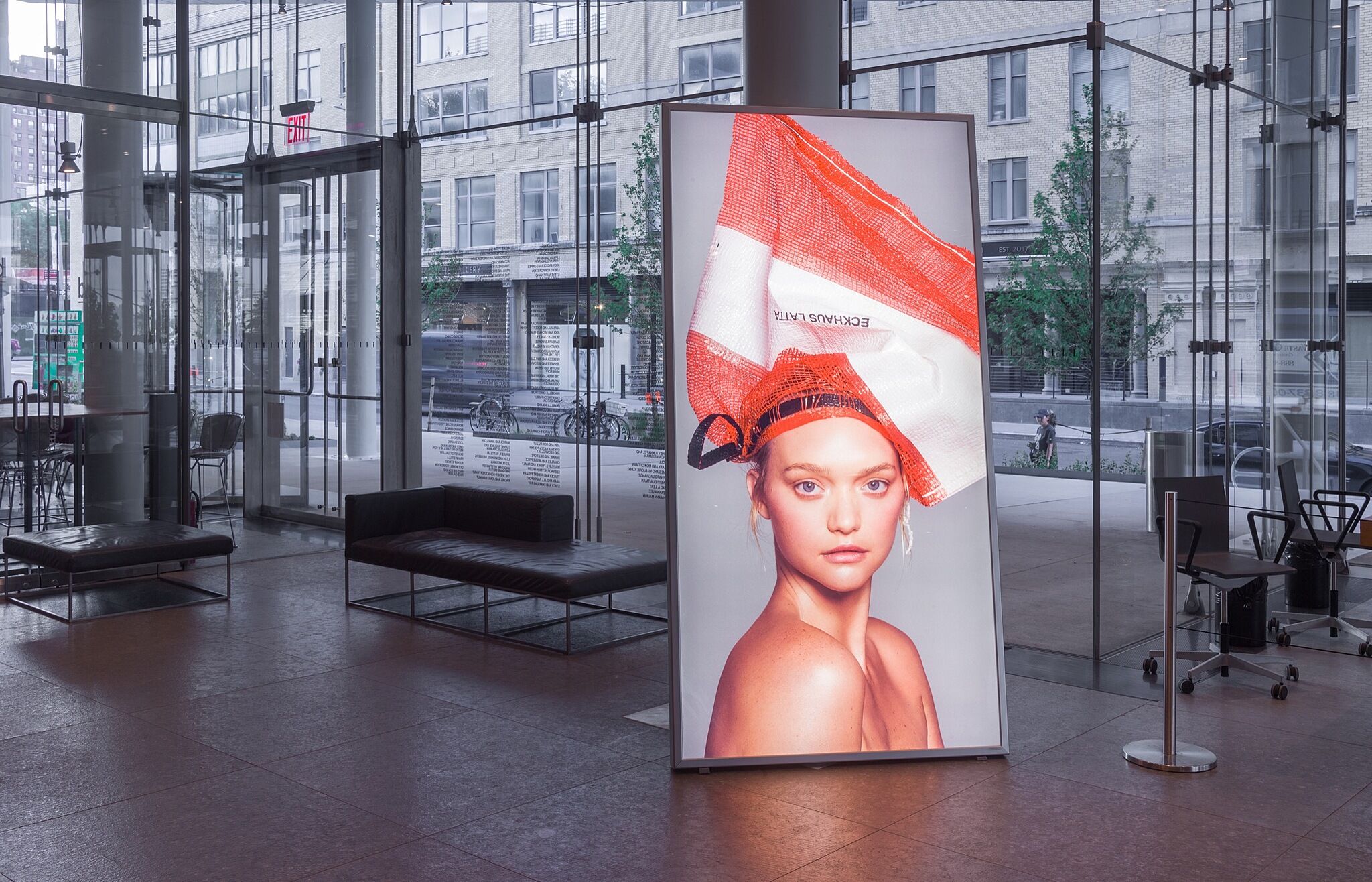 A lightbox photo of a blond model wearing a bag on their head in the Whitney lobby.