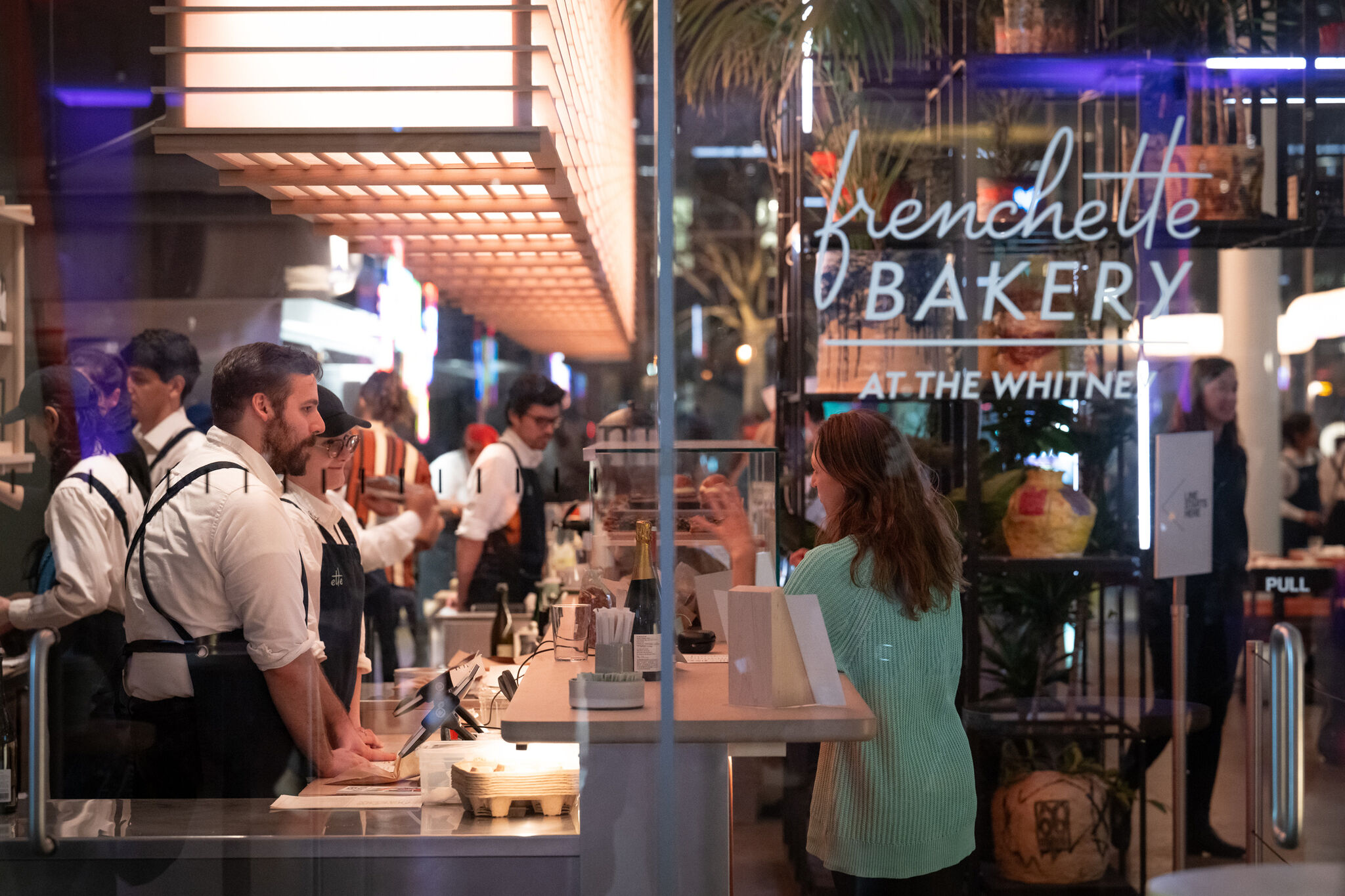 A lot of people seated in a time lapse, eating at a restaurant in front of a sign that says EAT, with bright lights and an orange wall. 