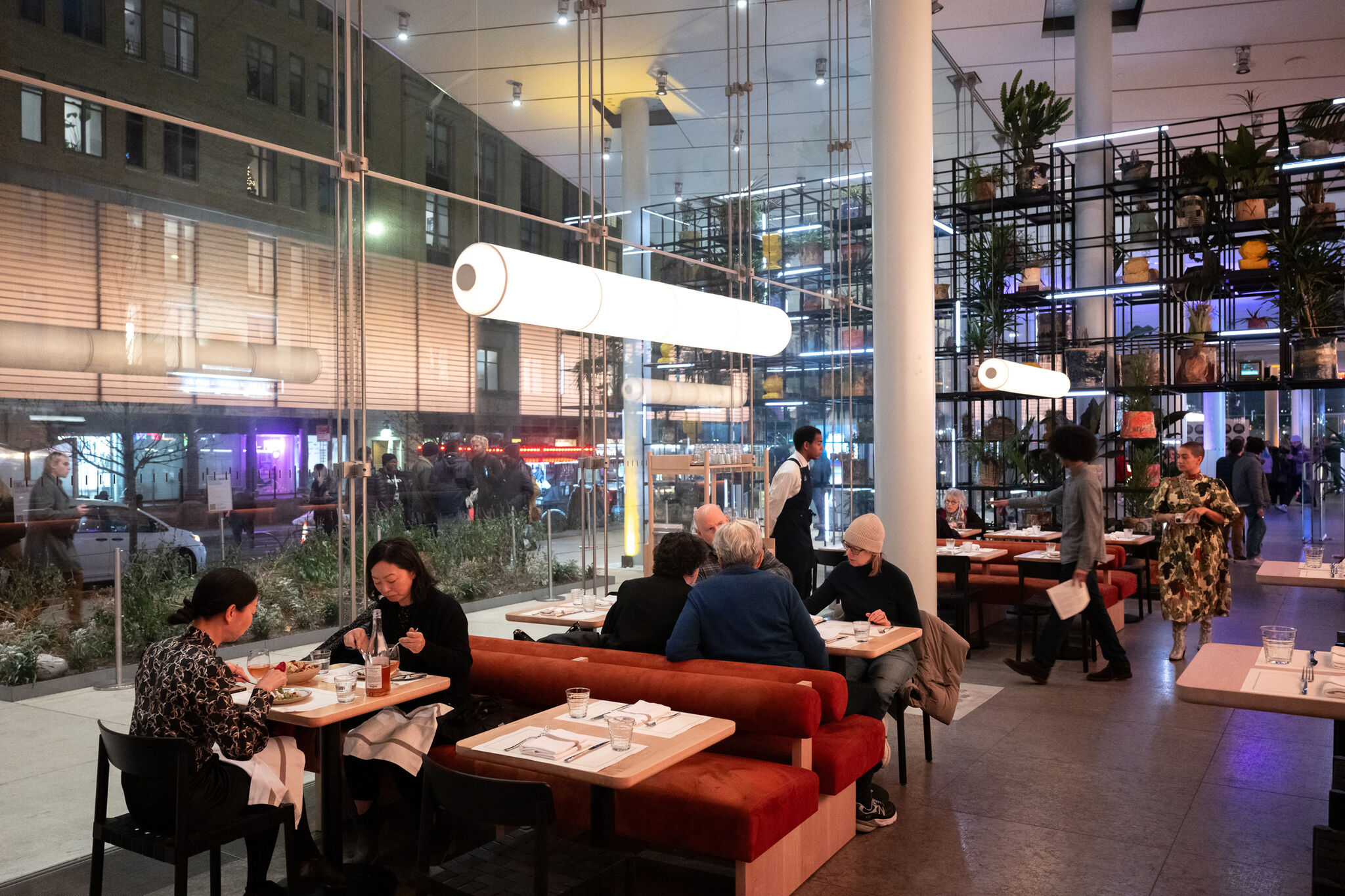 Modern restaurant interior with large windows, people dining at tables, and staff walking around. Shelves with plants and decor in the background.