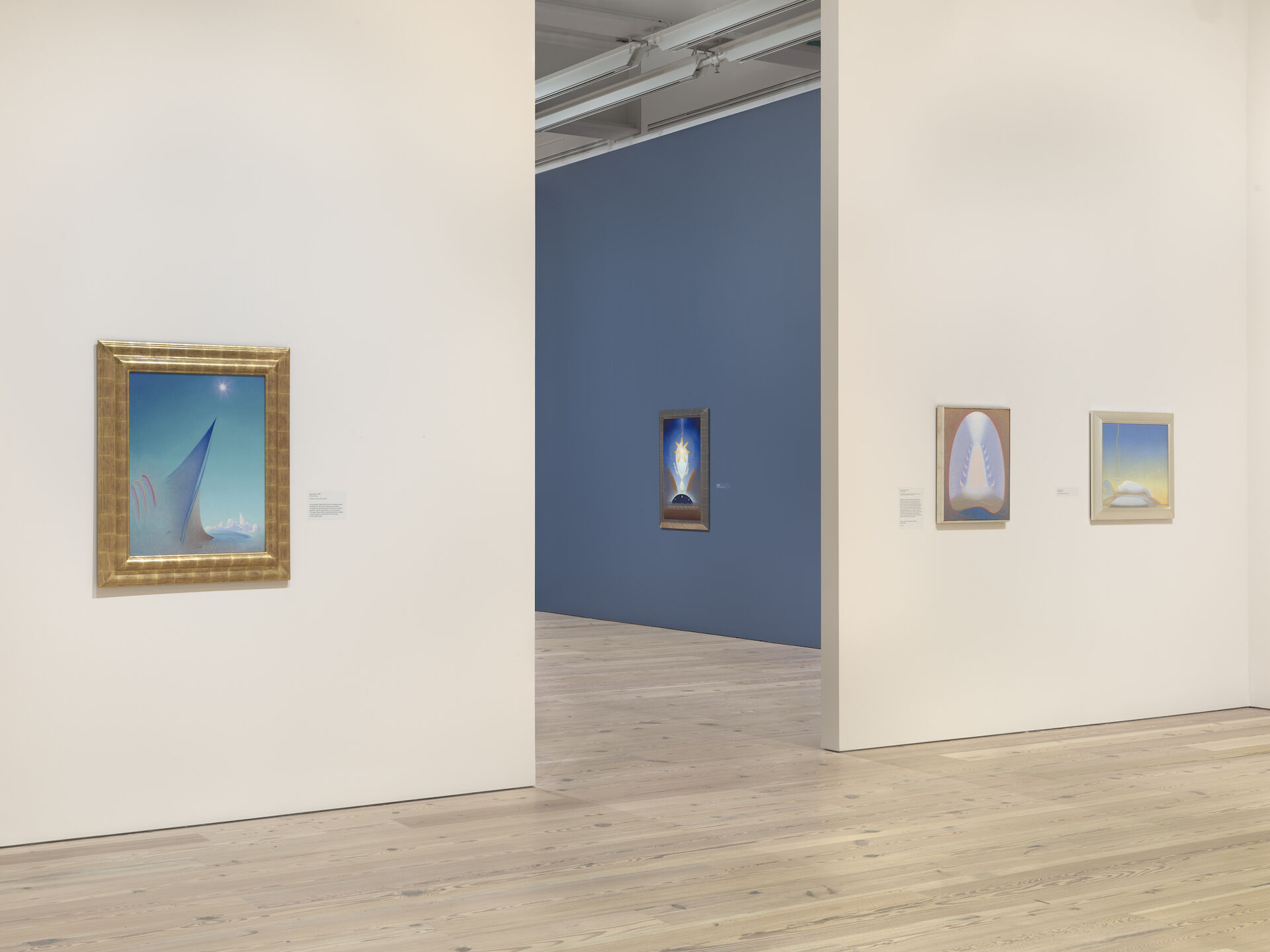 A white wall at the Whitney has three paintings surrounding a doorway to a blue room with more paintings from Agnes Pelton: Desert Transcendentalist at the Whitney.