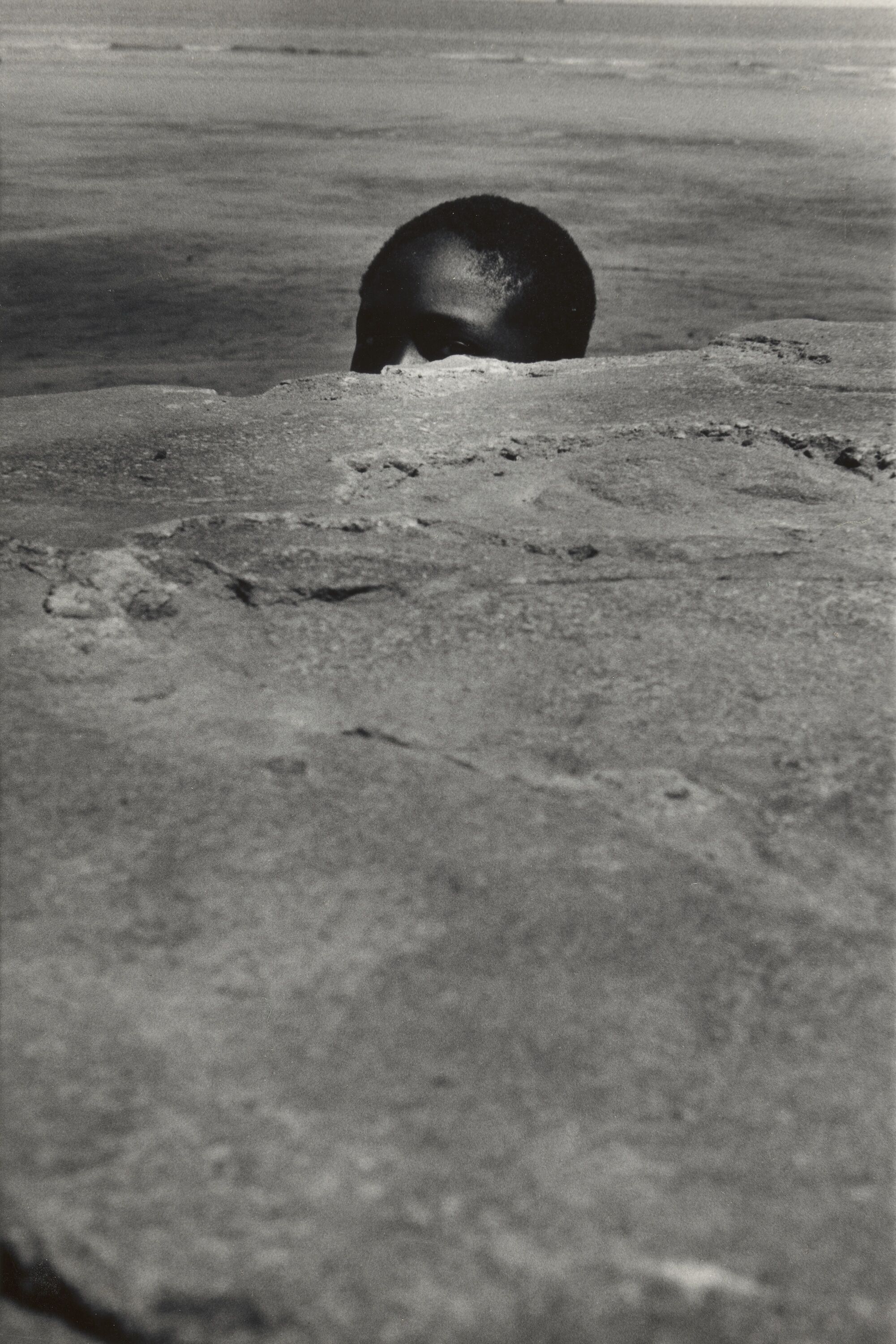 A child mostly hidden behind a rock formation. Only the top of their head is exposed above the rock plateau. 