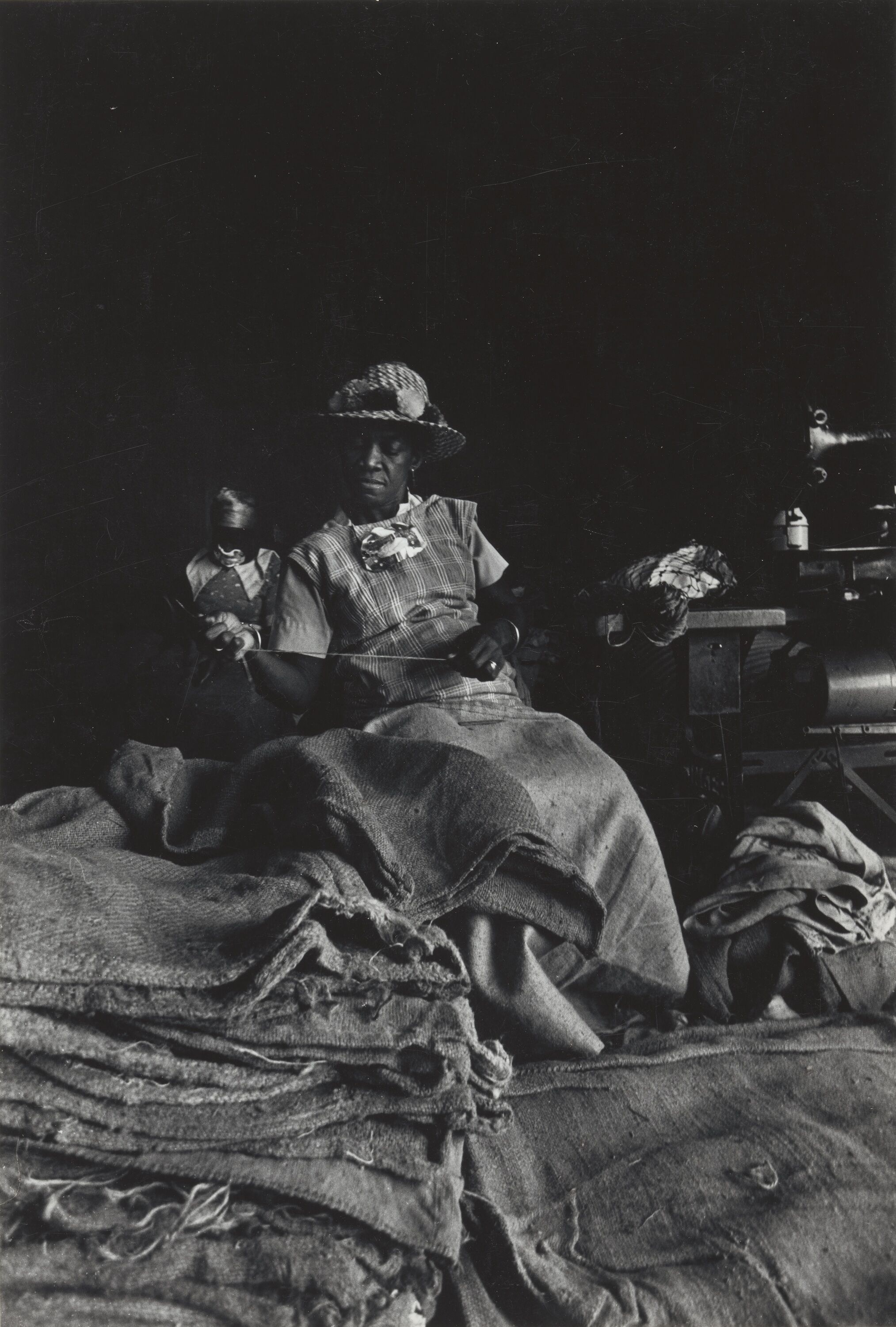A woman holding a strand of thread, sitting behind piles of empty bags. 