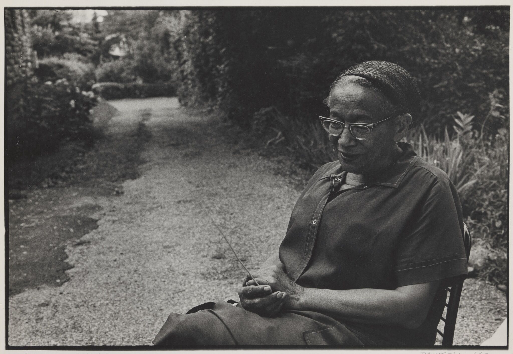 An elderly woman wearing glasses, sitting on a chair outside.