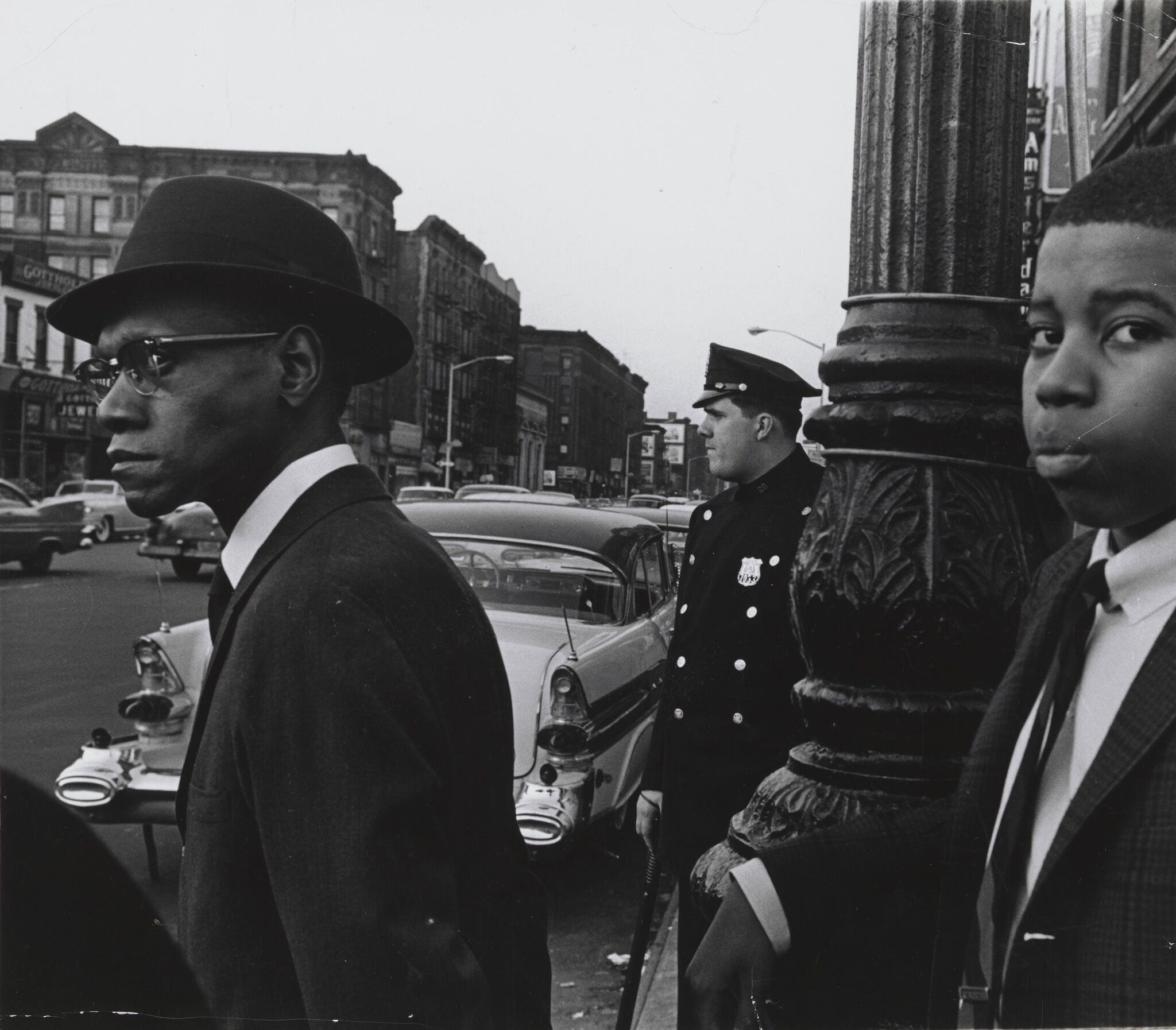 A black man and a young boy dressed in suits standing outside on a street with a white man in uniform standing nearby.