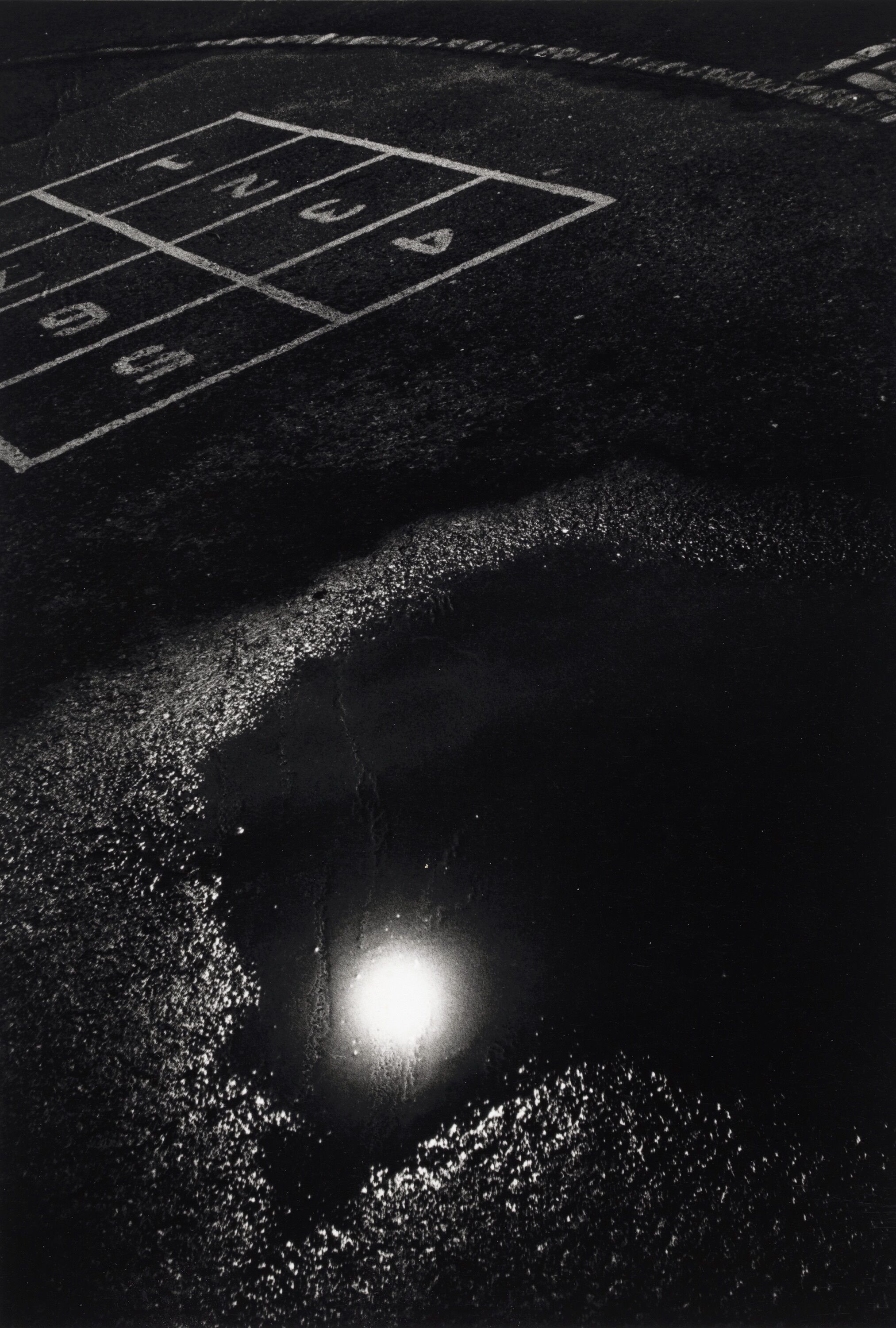 A dark pavement with a large puddle reflecting a source of light.