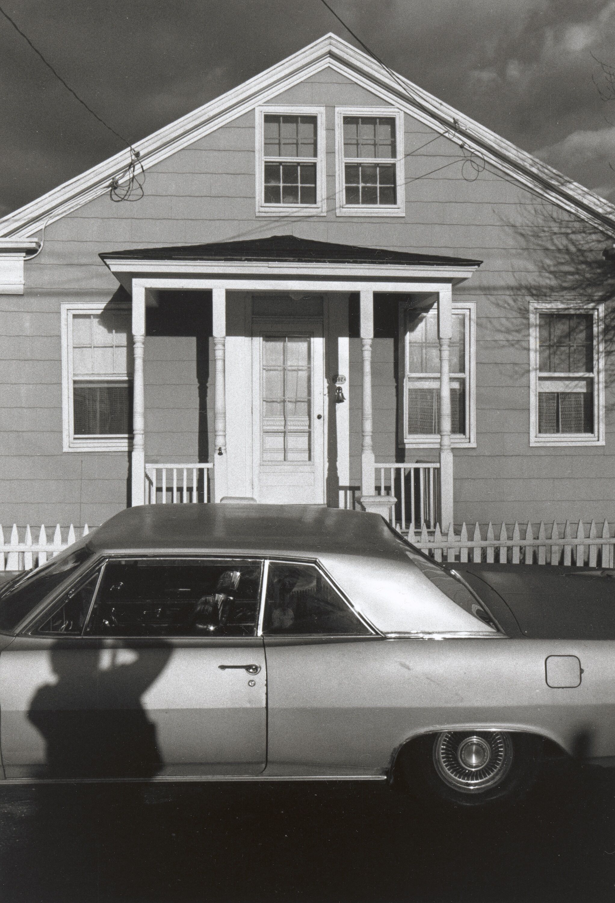 A house with a car parked in front of it, along with a shadow of a person shown on the car.